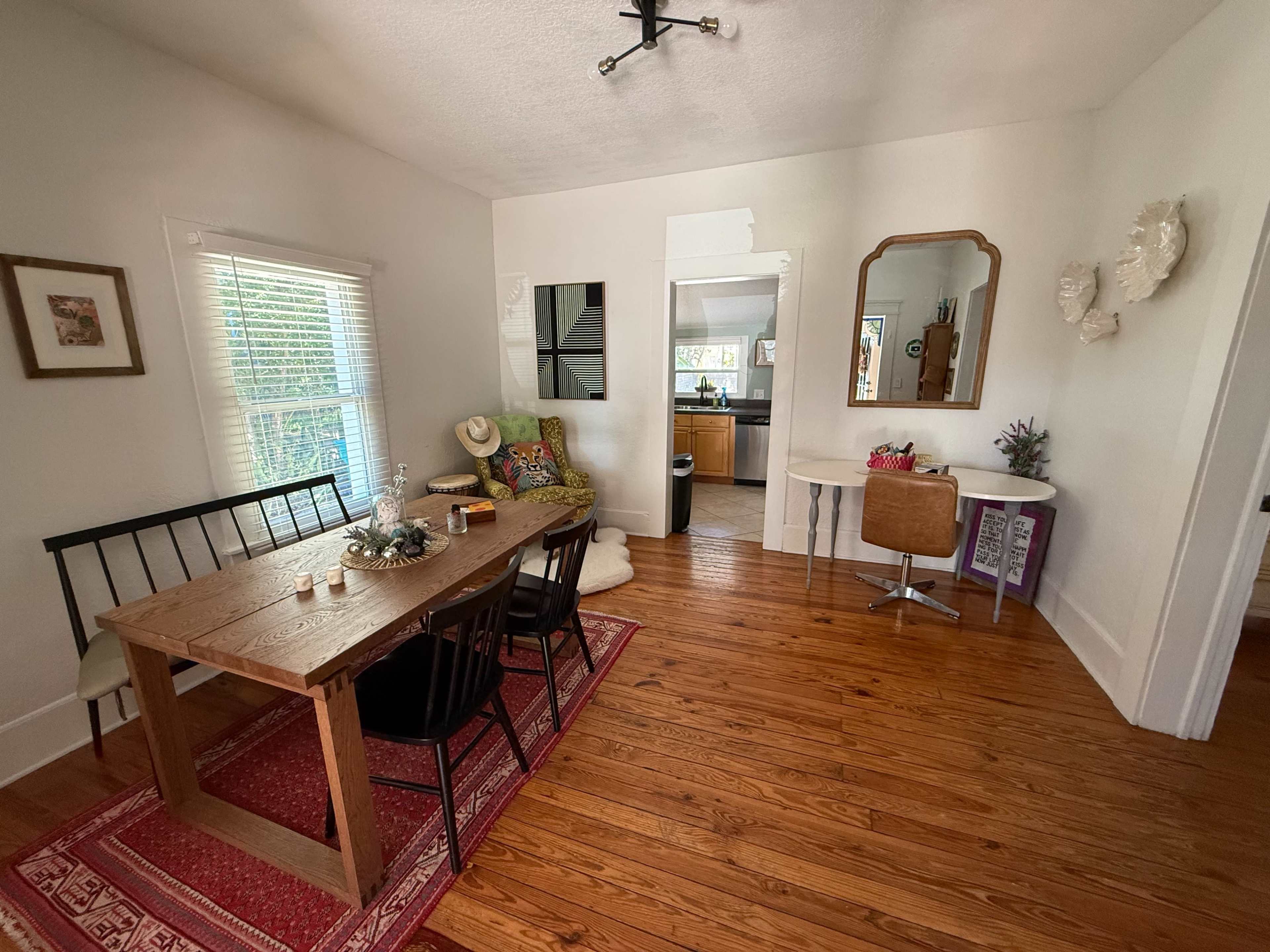 The image shows a cozy dining area with a wooden table and black chairs, framed by light-colored walls and hardwood floors, leading to a doorway that opens into a kitchen space.