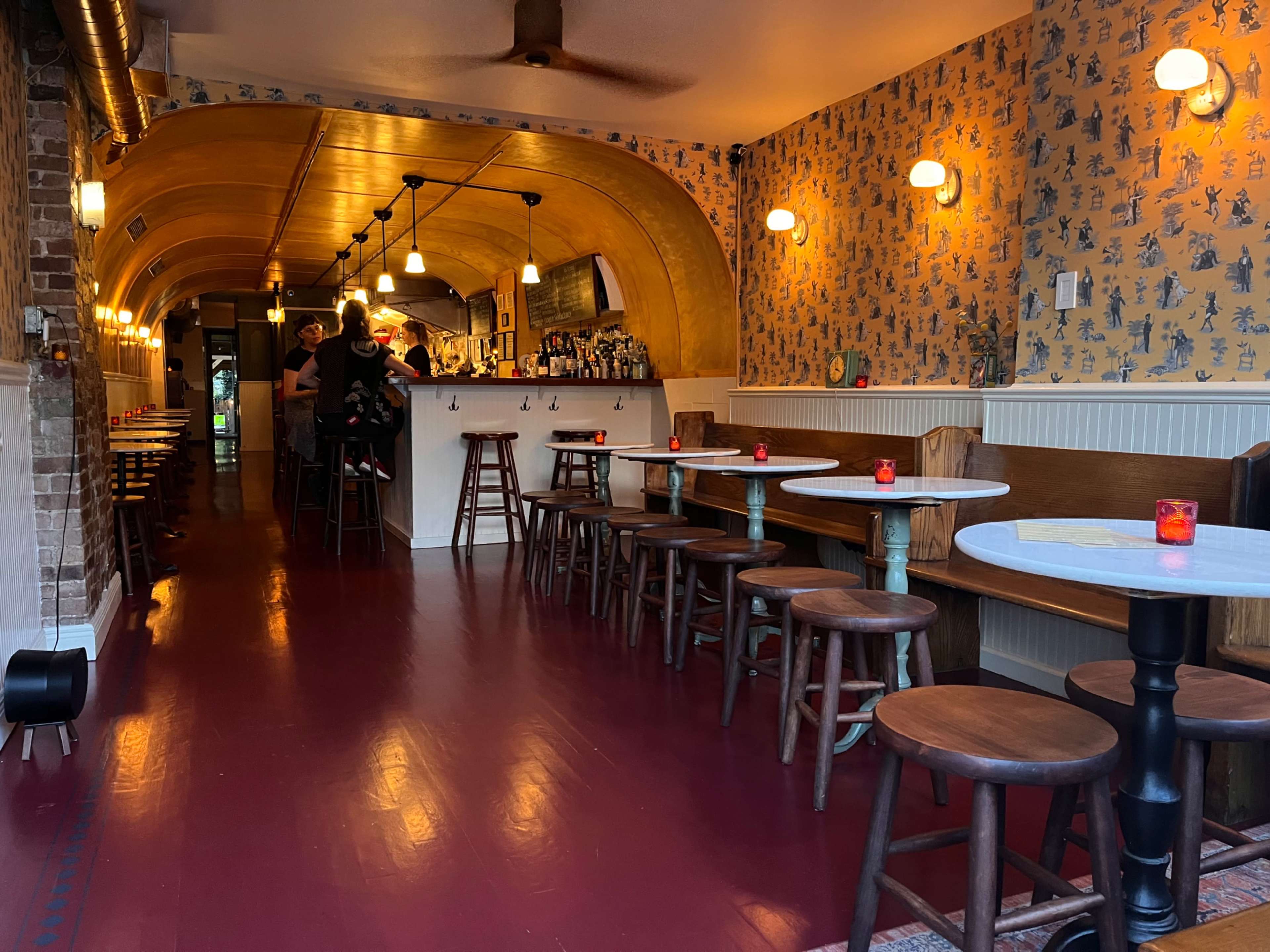 The image shows a narrow restaurant interior with wooden stools and tables, featuring a bar area at the back and decorative wallpaper.