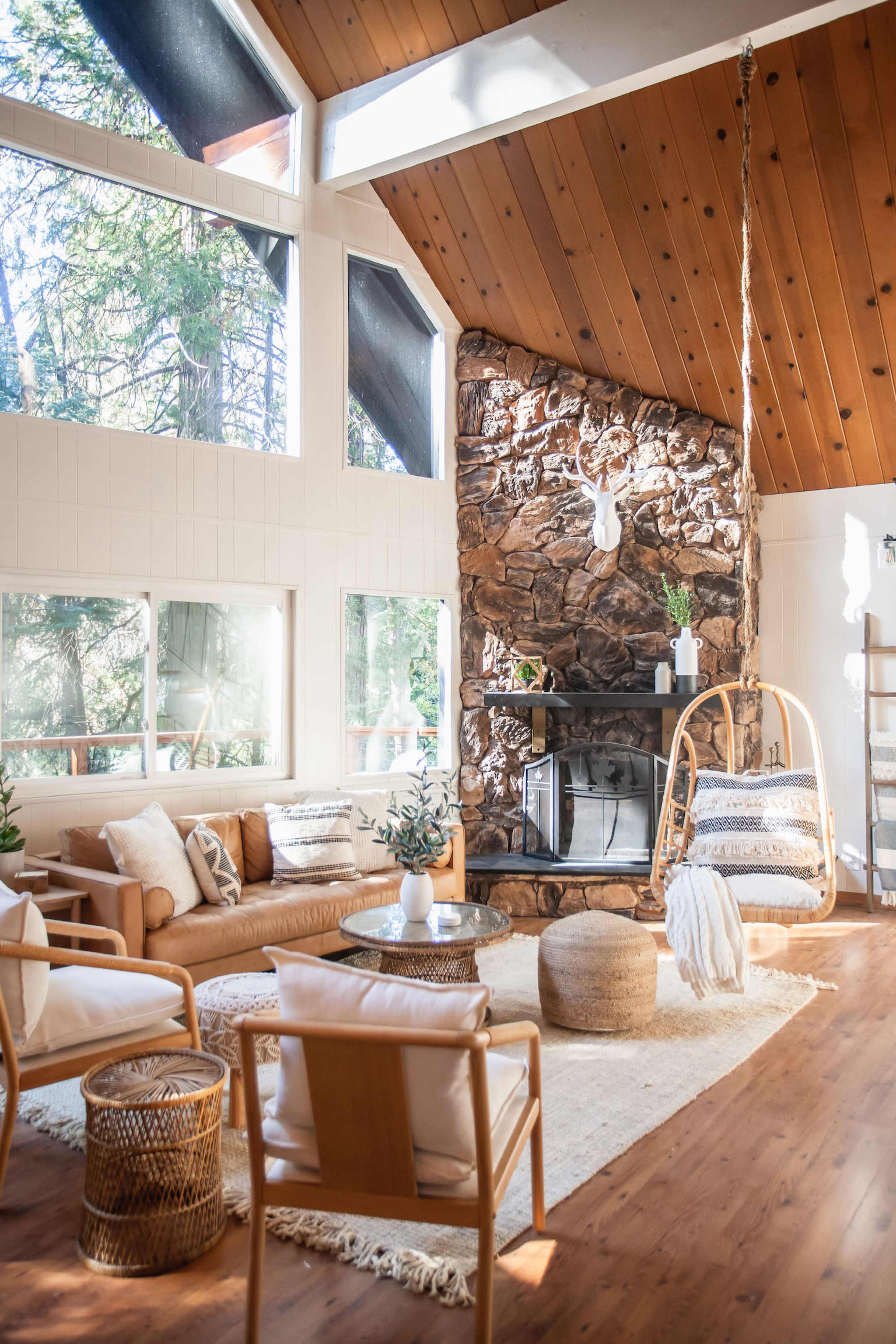 The image shows a modern living room with large windows, a stone fireplace, wooden beams, and a hanging chair.