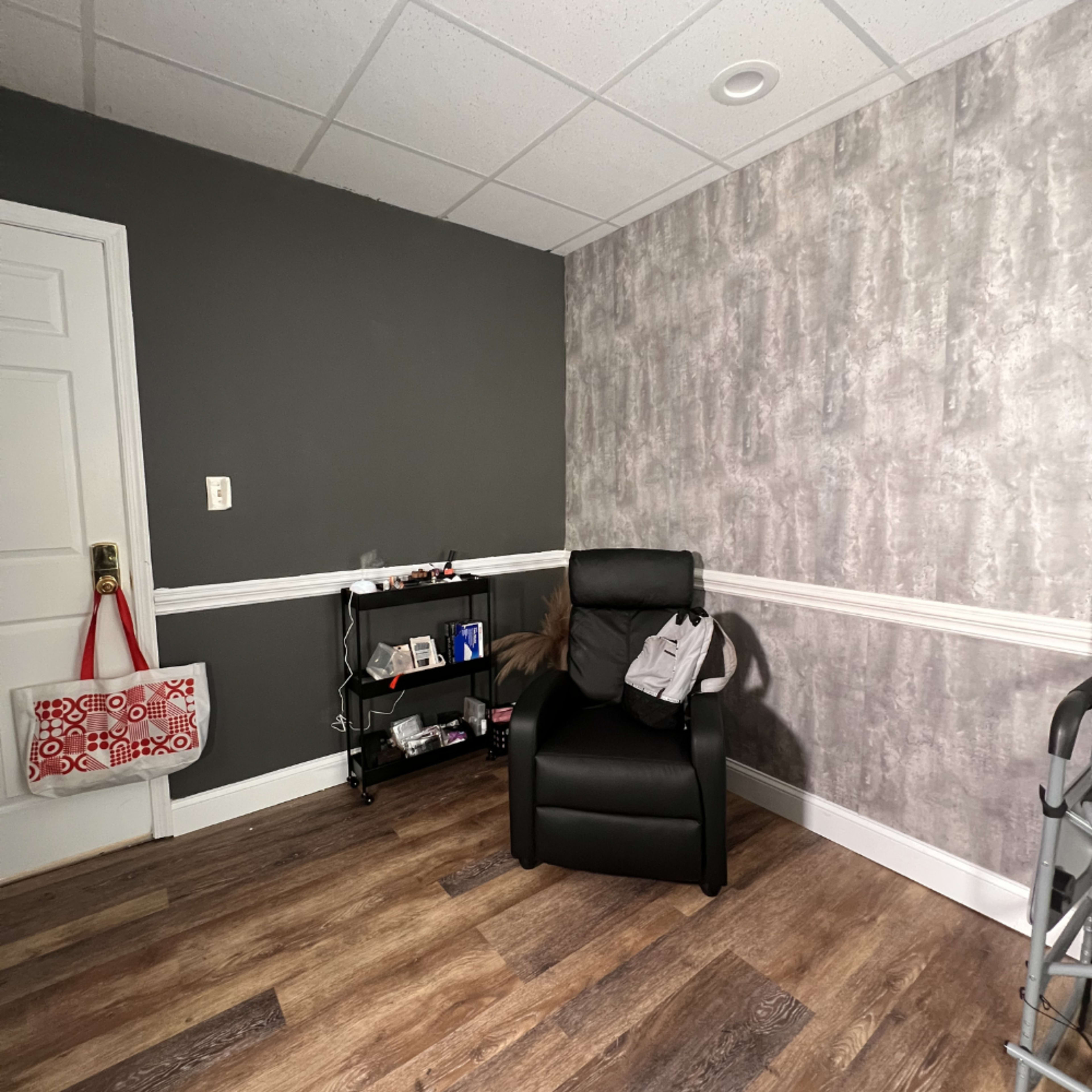 The image shows a small room featuring a black chair, a shelf filled with various items, a gray wall, and a patterned wall opposite the chair.