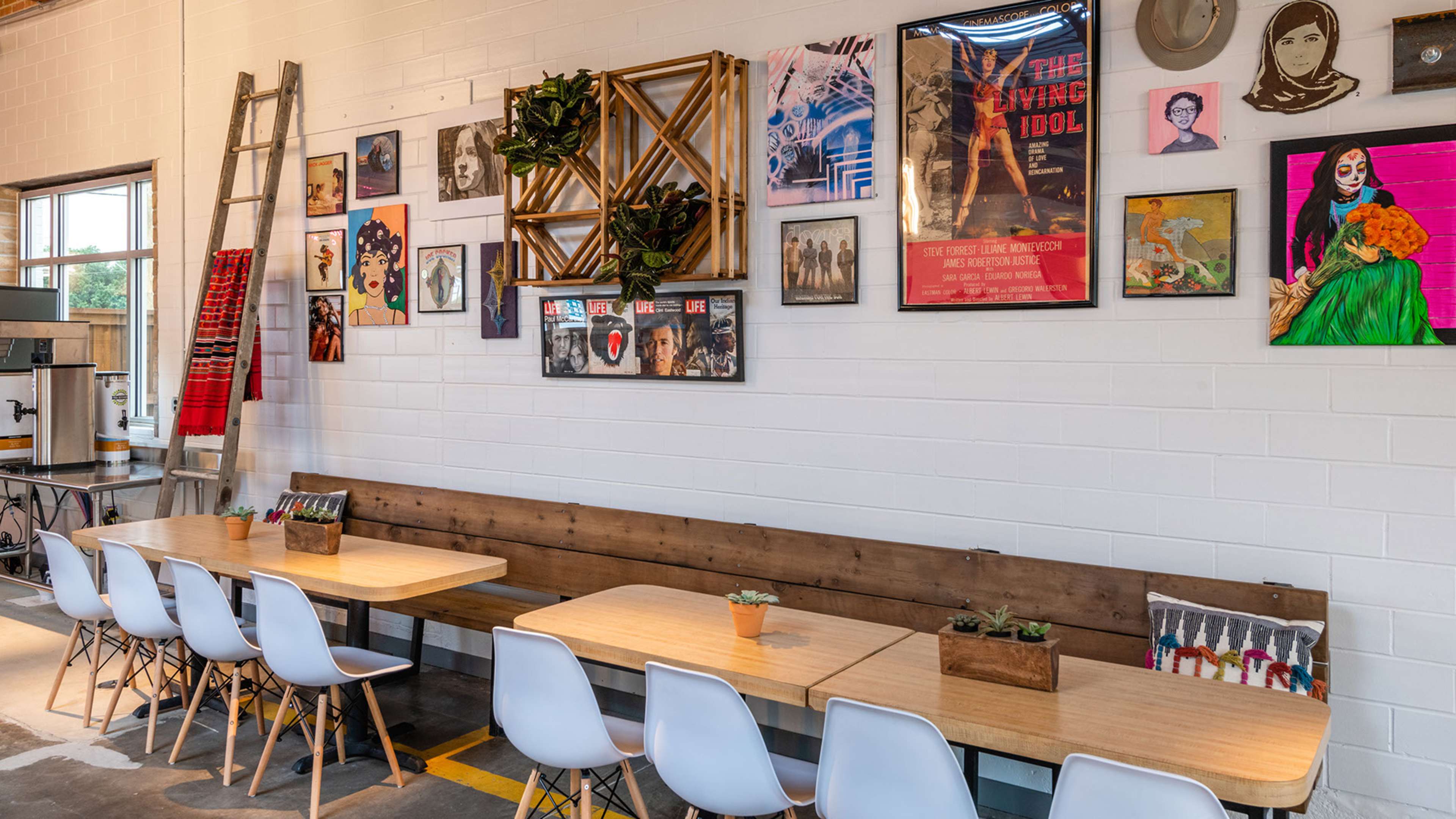 A modern cafe interior with wooden tables and white chairs arranged along a wall adorned with various artworks and framed posters.