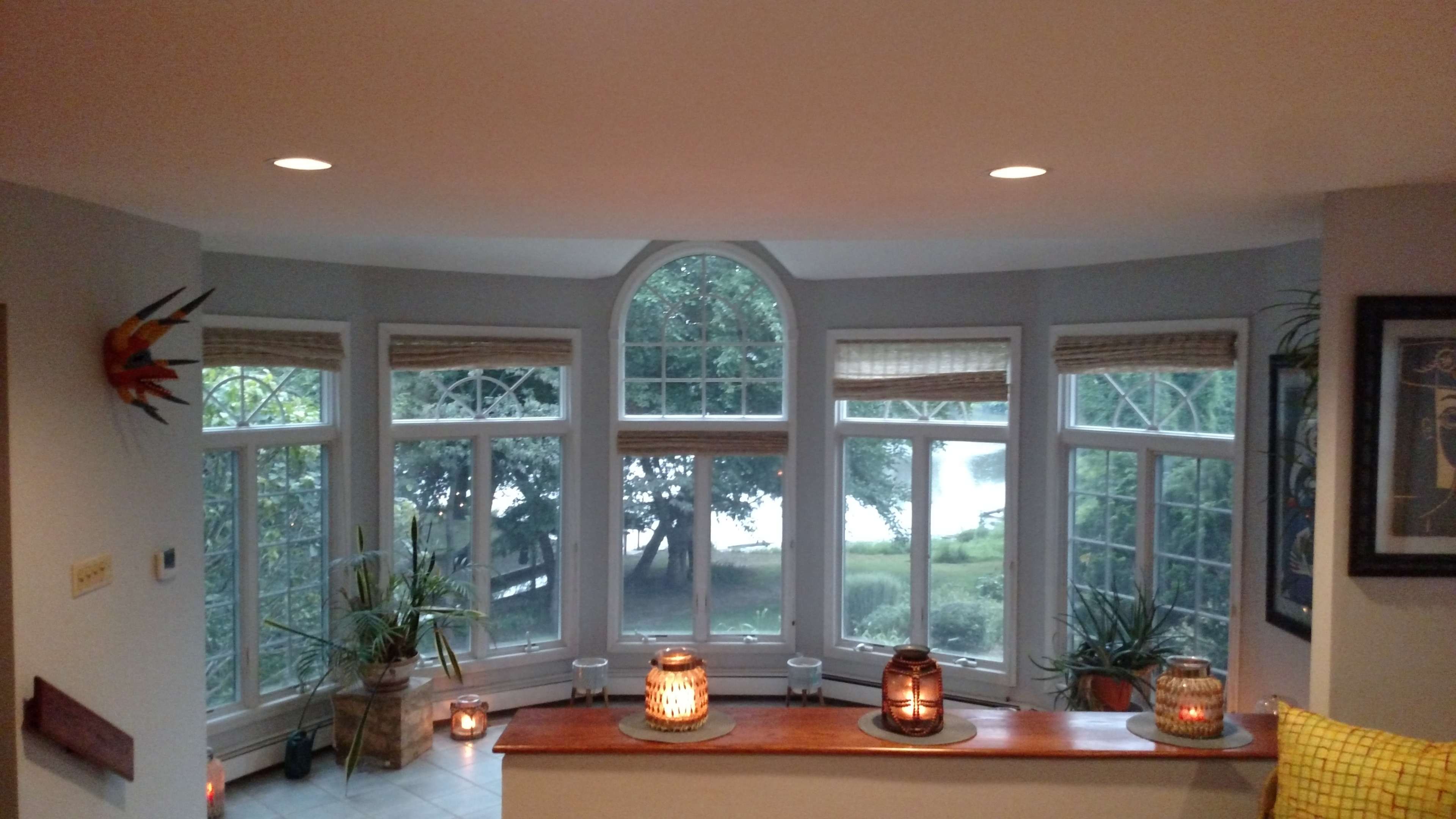 The image shows a bright room with large bay windows overlooking a scenic view, adorned with plants and candles on a ledge.