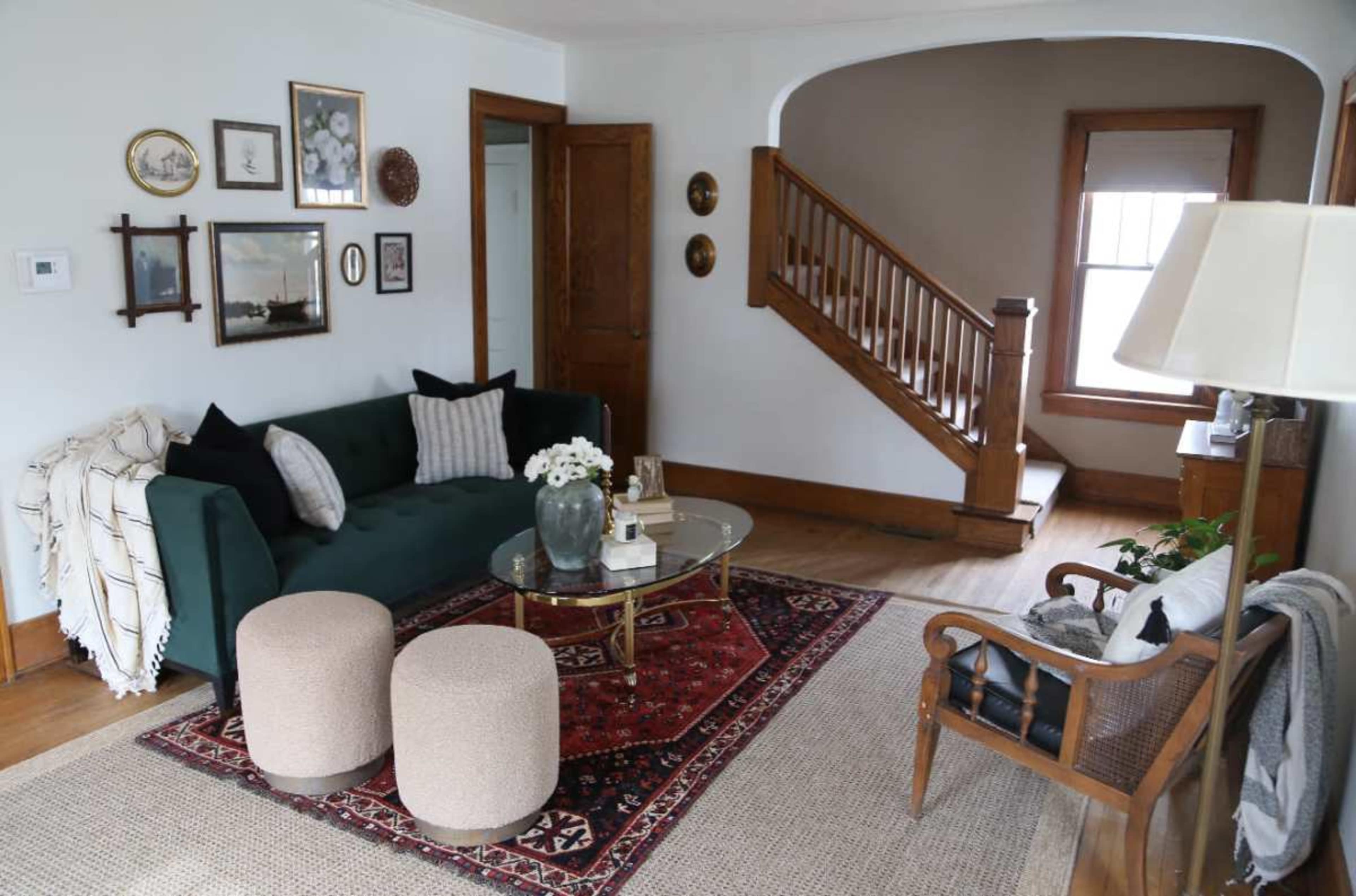 The image shows a cozy living room with a green sofa, two round, beige ottomans, a glass coffee table, and a staircase in the background.