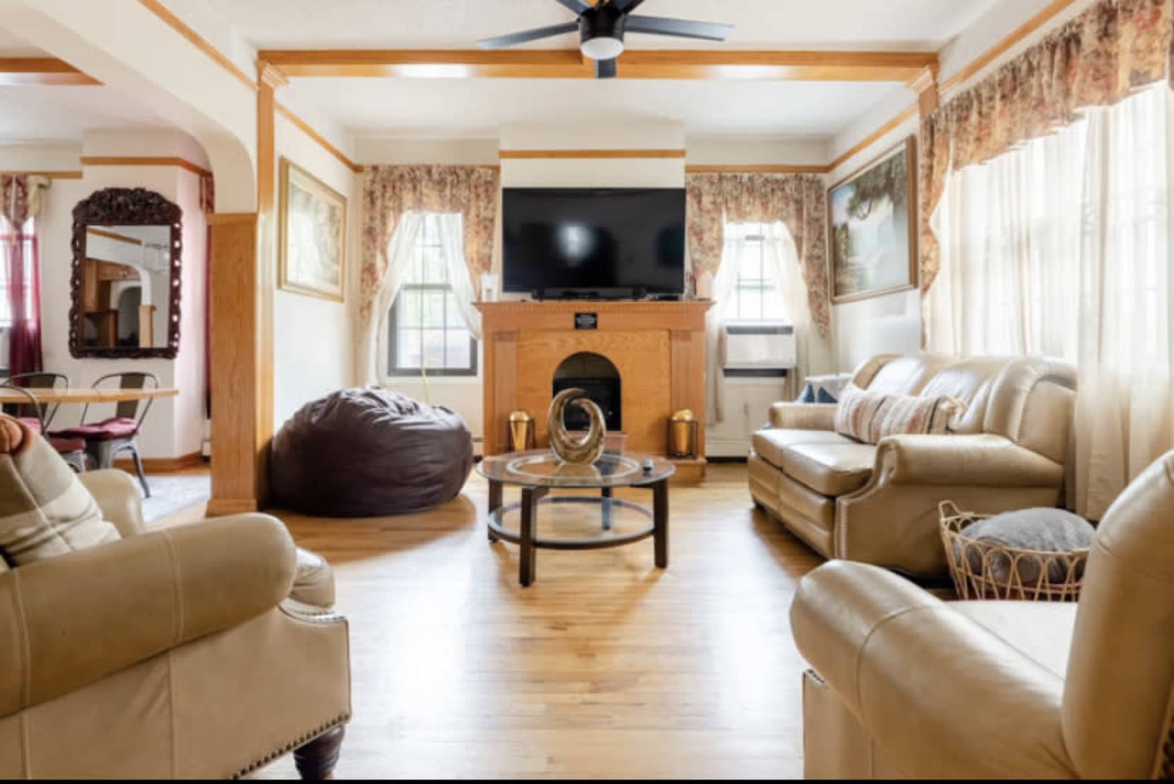 A living room with beige leather furniture, a wooden fireplace, a large television mounted on the wall, and a round coffee table in the center.
