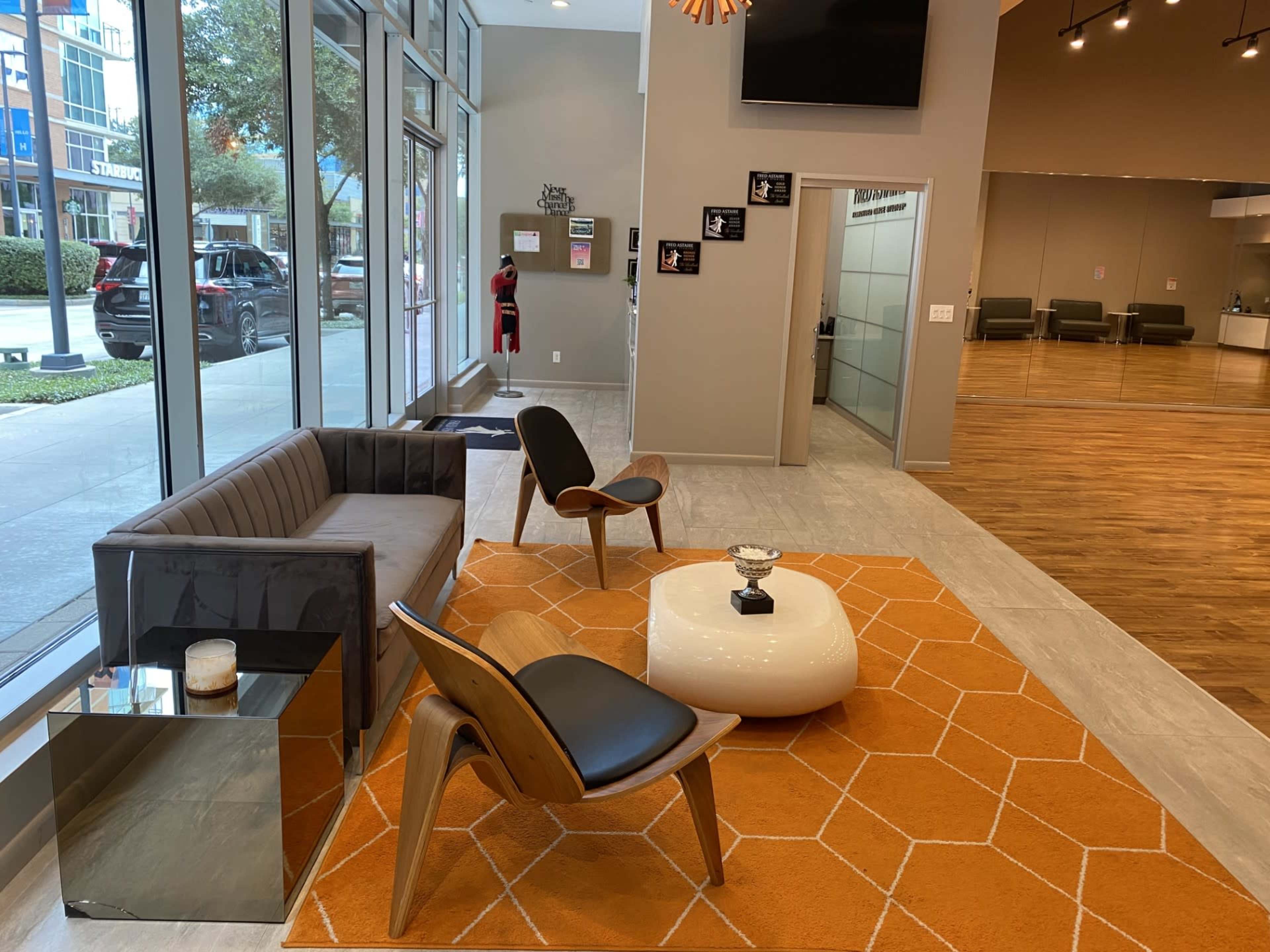 The image shows a modern reception area with a gray sofa, wooden chairs, an orange geometric rug, and large windows letting in natural light.