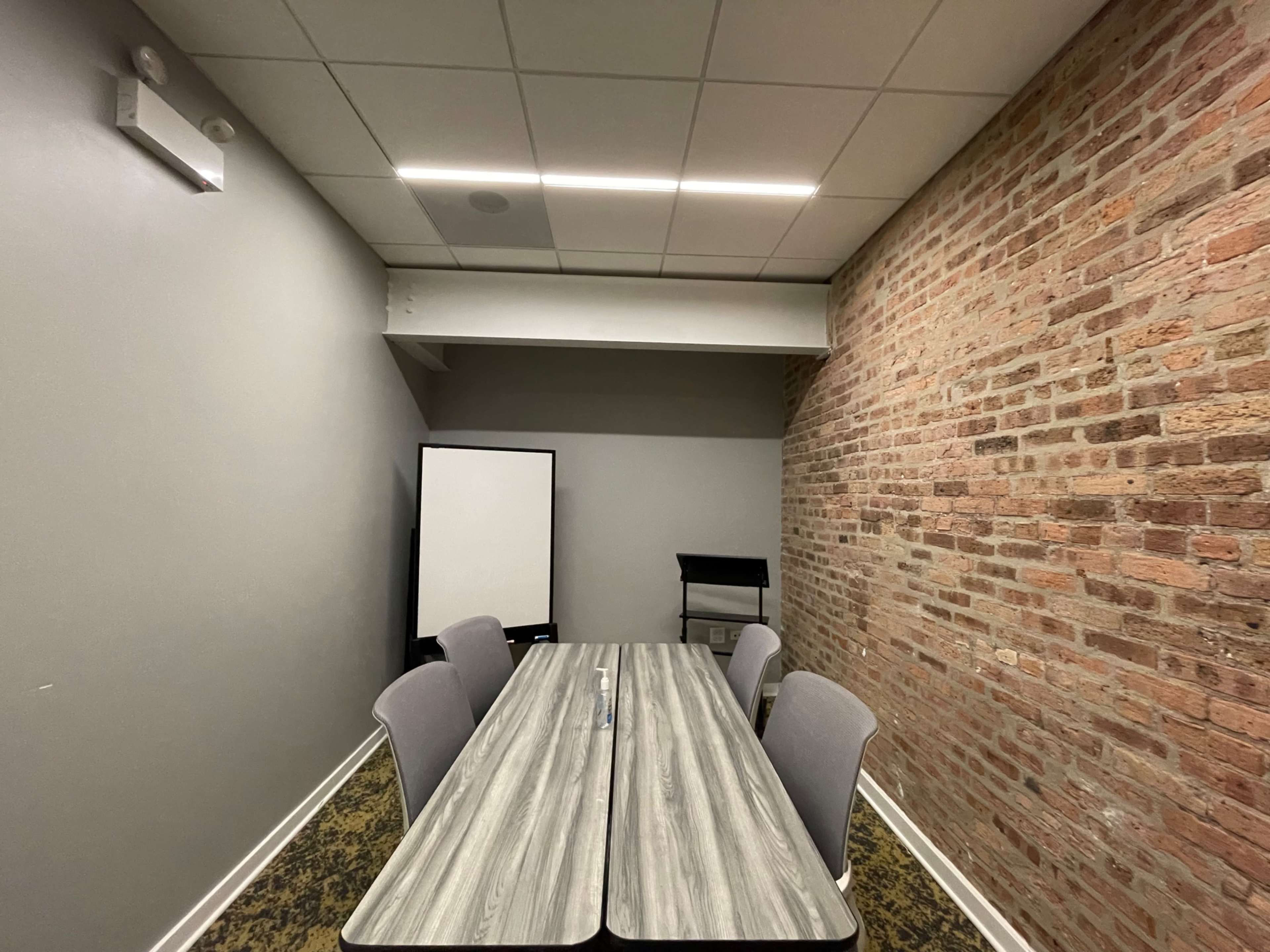 A small conference room features a rectangular table surrounded by four chairs, with a brick wall on one side and a projector screen at the back.
