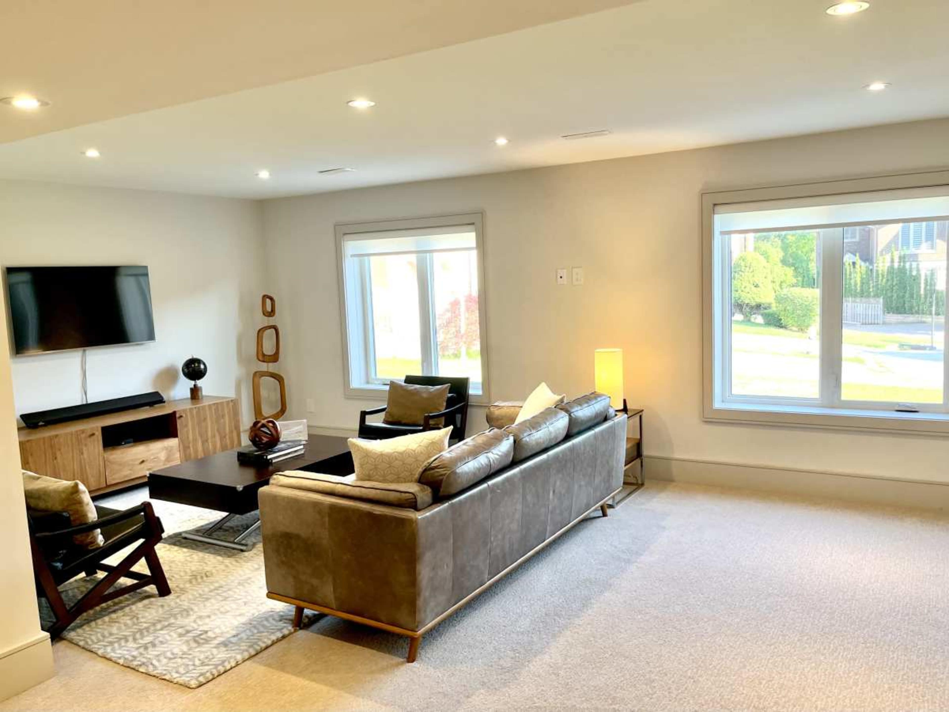 The image shows a modern living room with a television unit, a sofa, armchairs, and large windows letting in natural light.