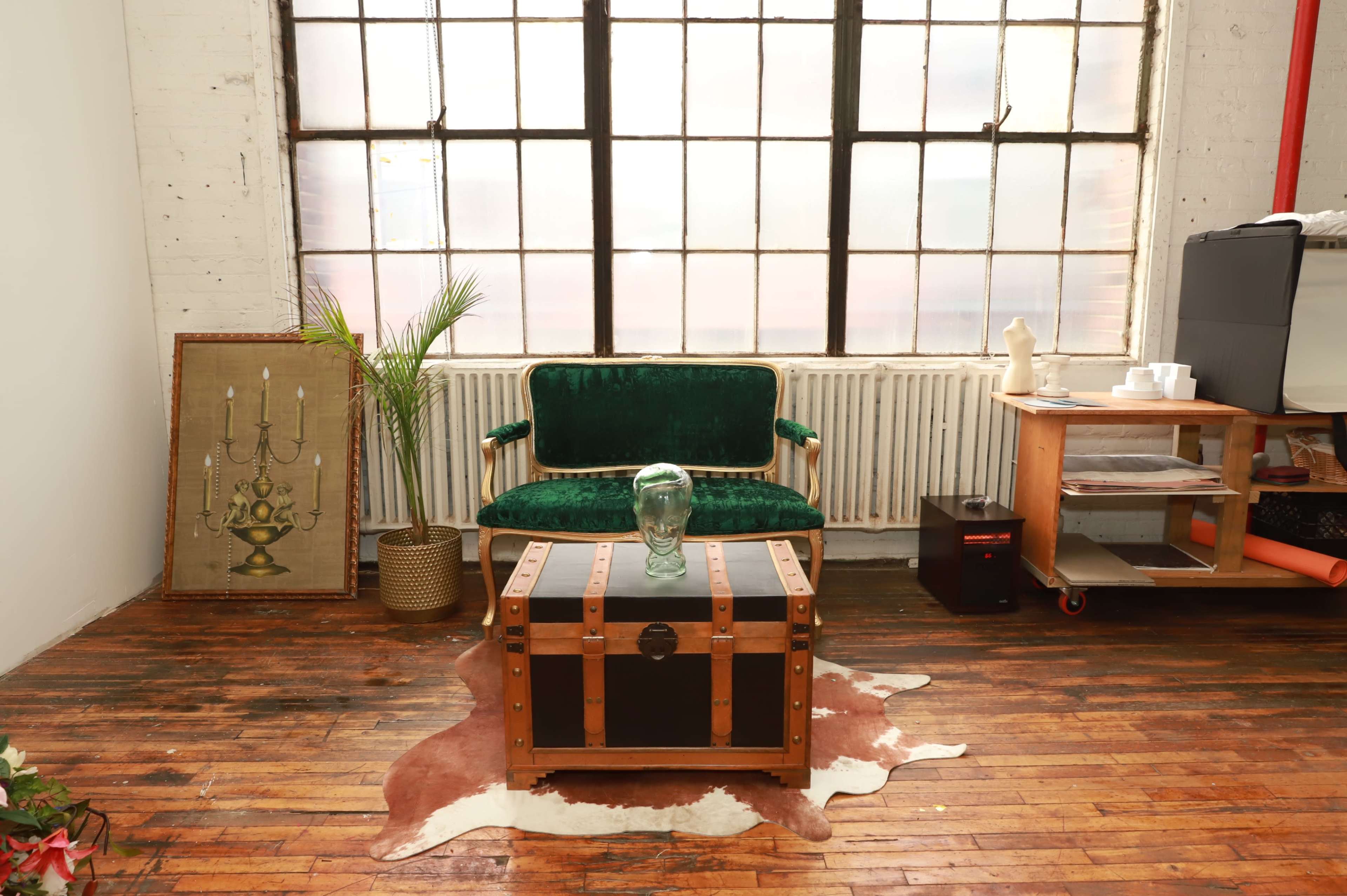 A green velvet sofa is positioned in front of a large window, with a wooden trunk and a decorative vase on a table in a rustic room with wooden floors.