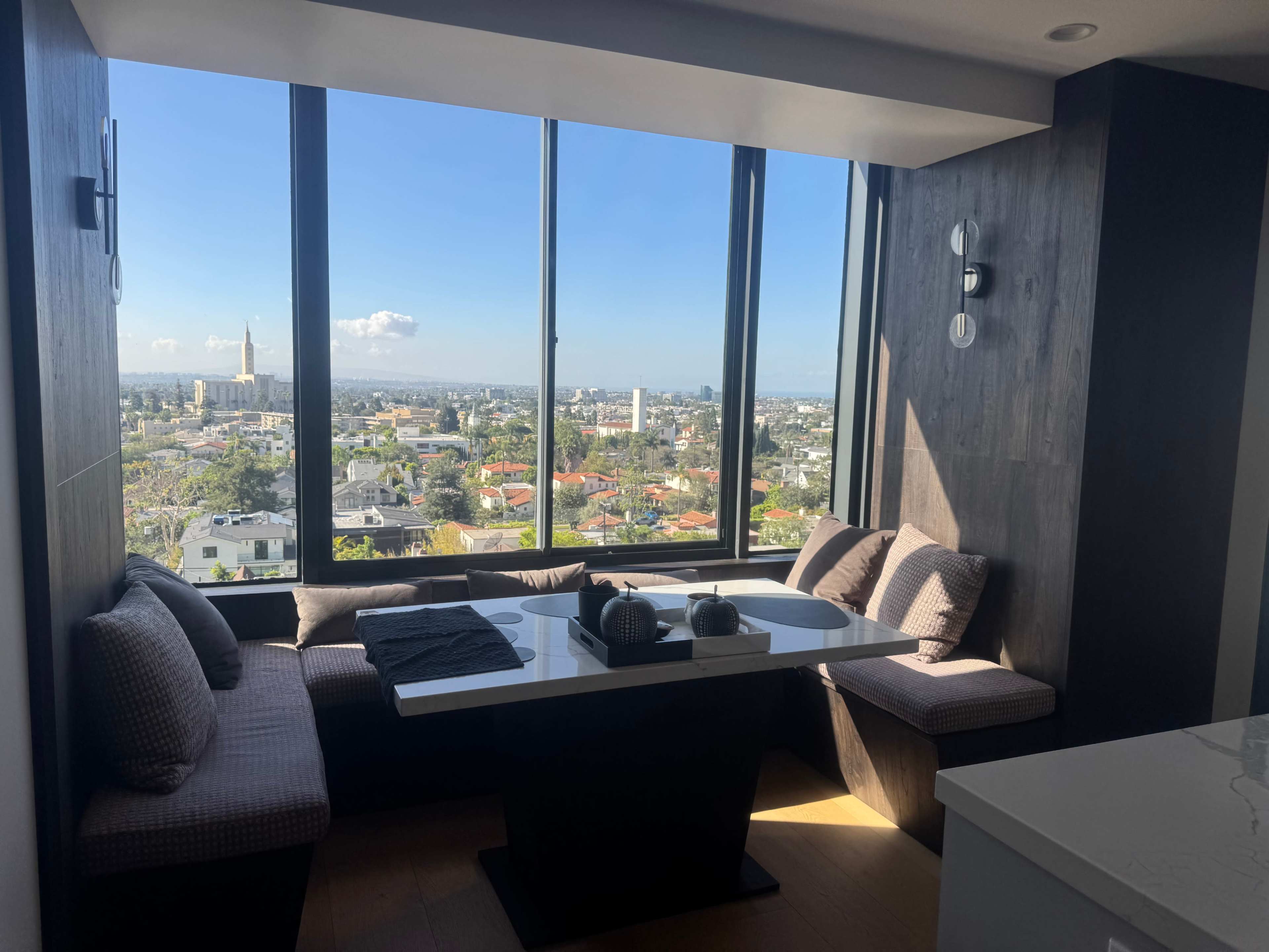 A modern dining nook with a table and cushioned seating offers a view of a city skyline through large windows.