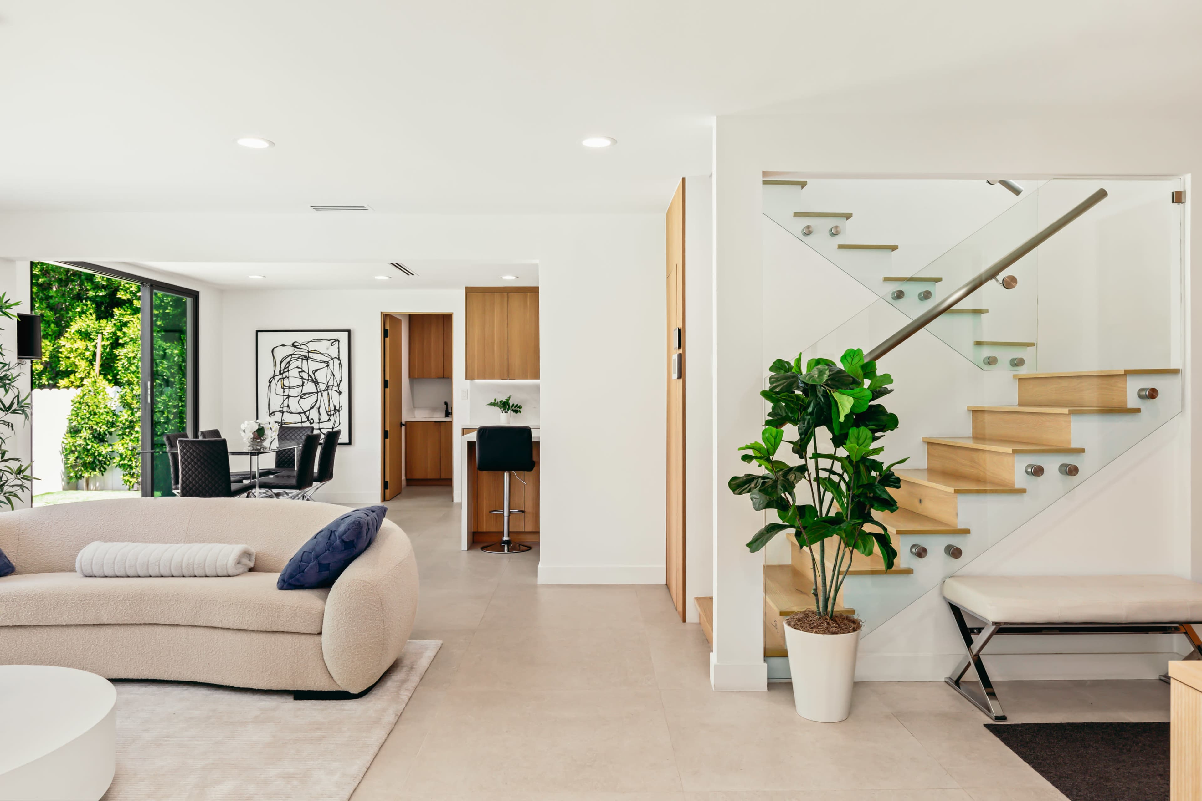 The image shows a modern interior space featuring a beige curved sofa, a glass staircase, and an open kitchen area visible in the background.