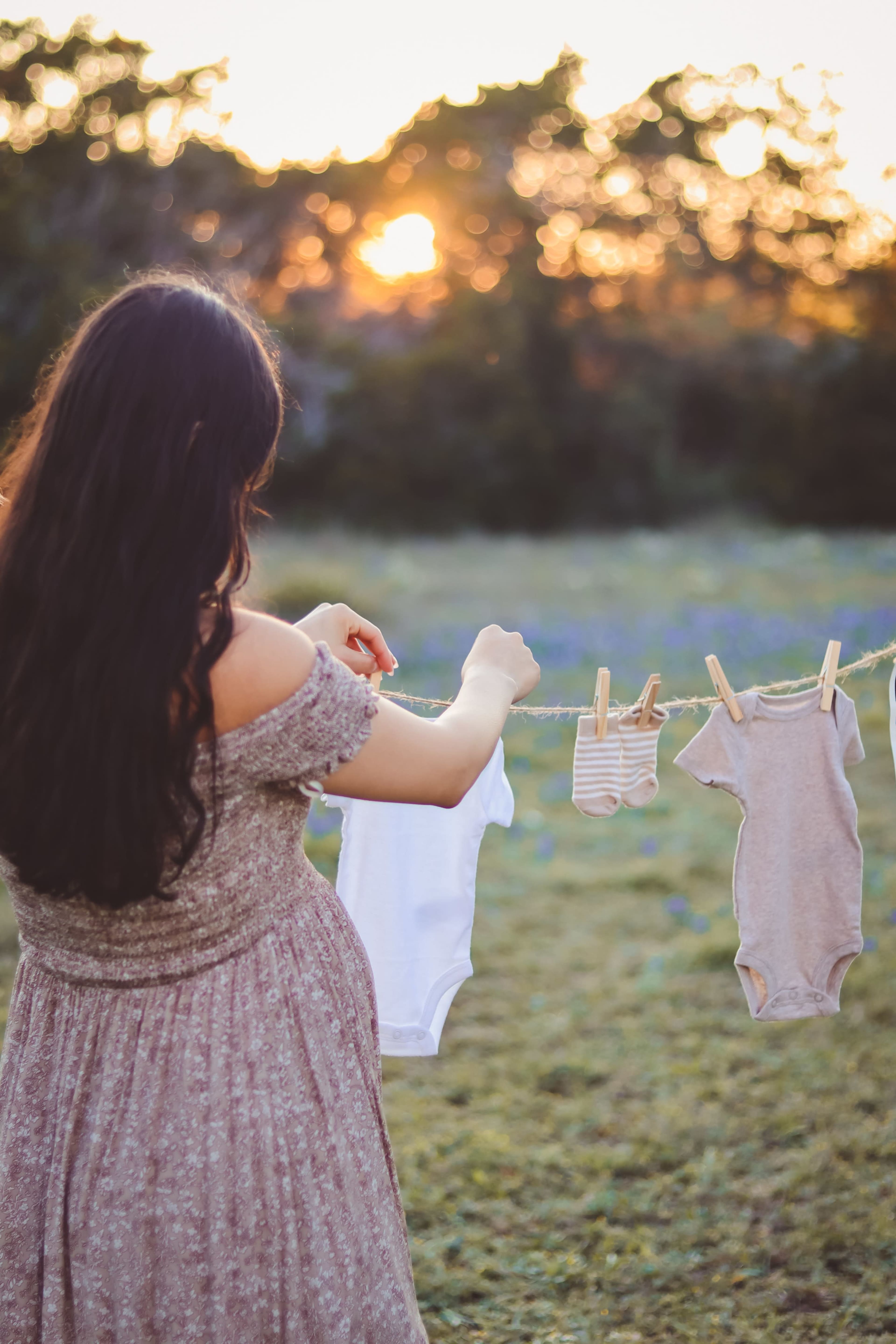 A woman stands in a field at sunset, hanging baby clothes on a clothesline.