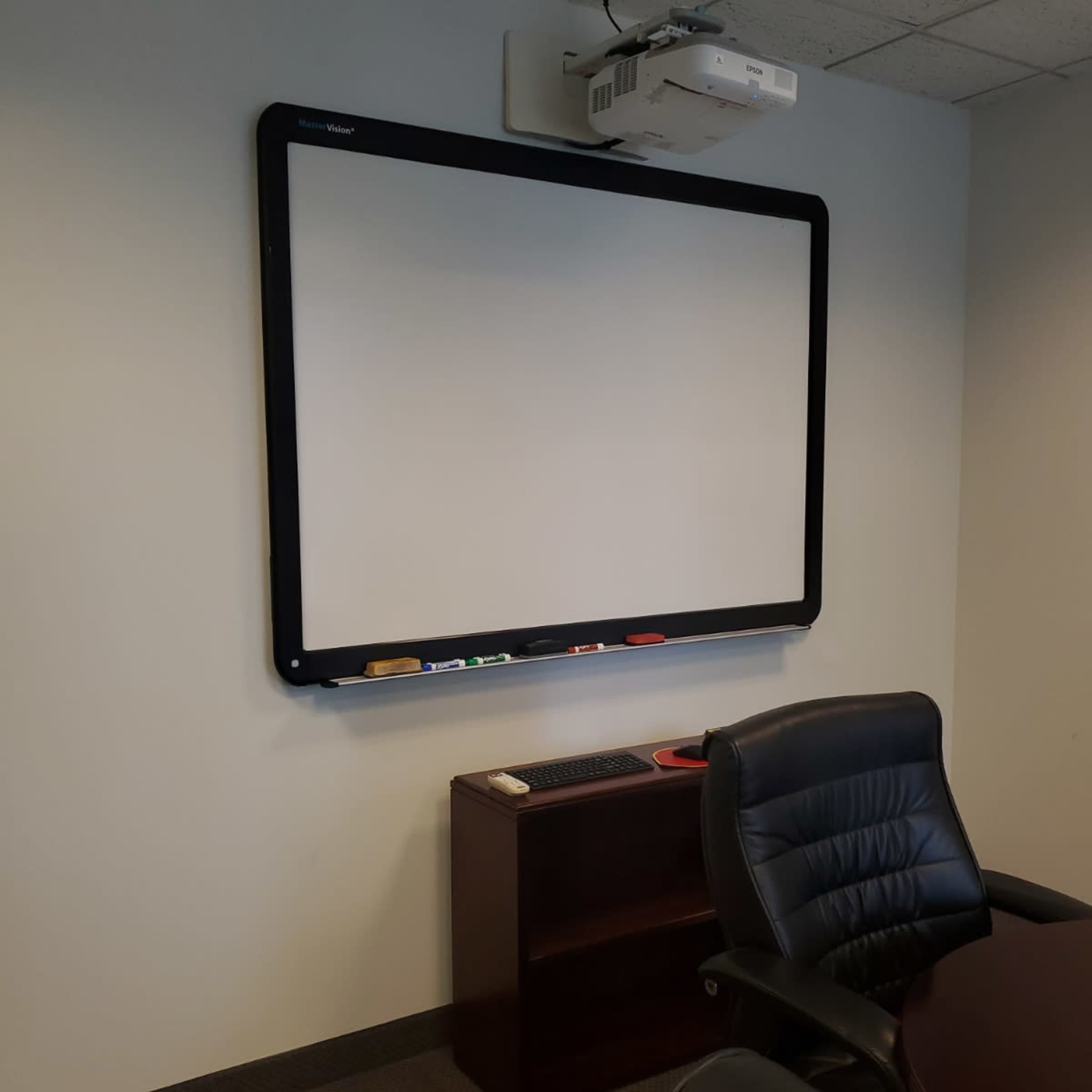 A large blank whiteboard mounted on a wall above a dark wooden desk and a black ergonomic chair in an office setting.