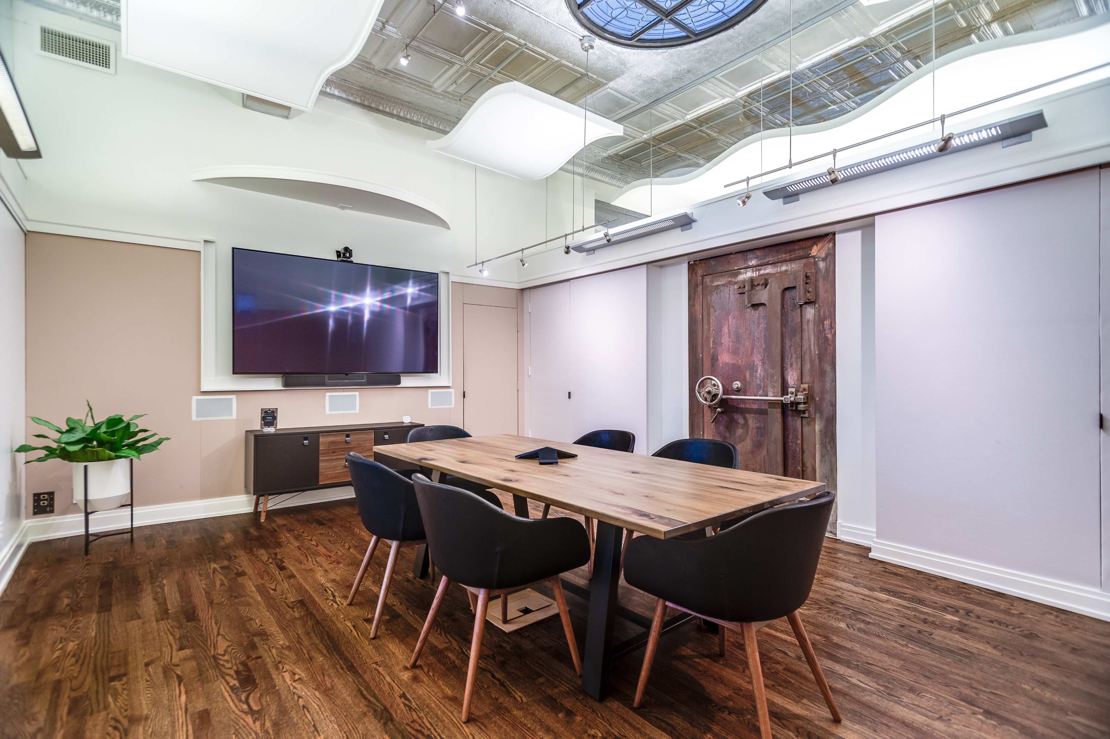 A modern conference room features a wooden table surrounded by black chairs, a wall-mounted television, and a large decorative door.