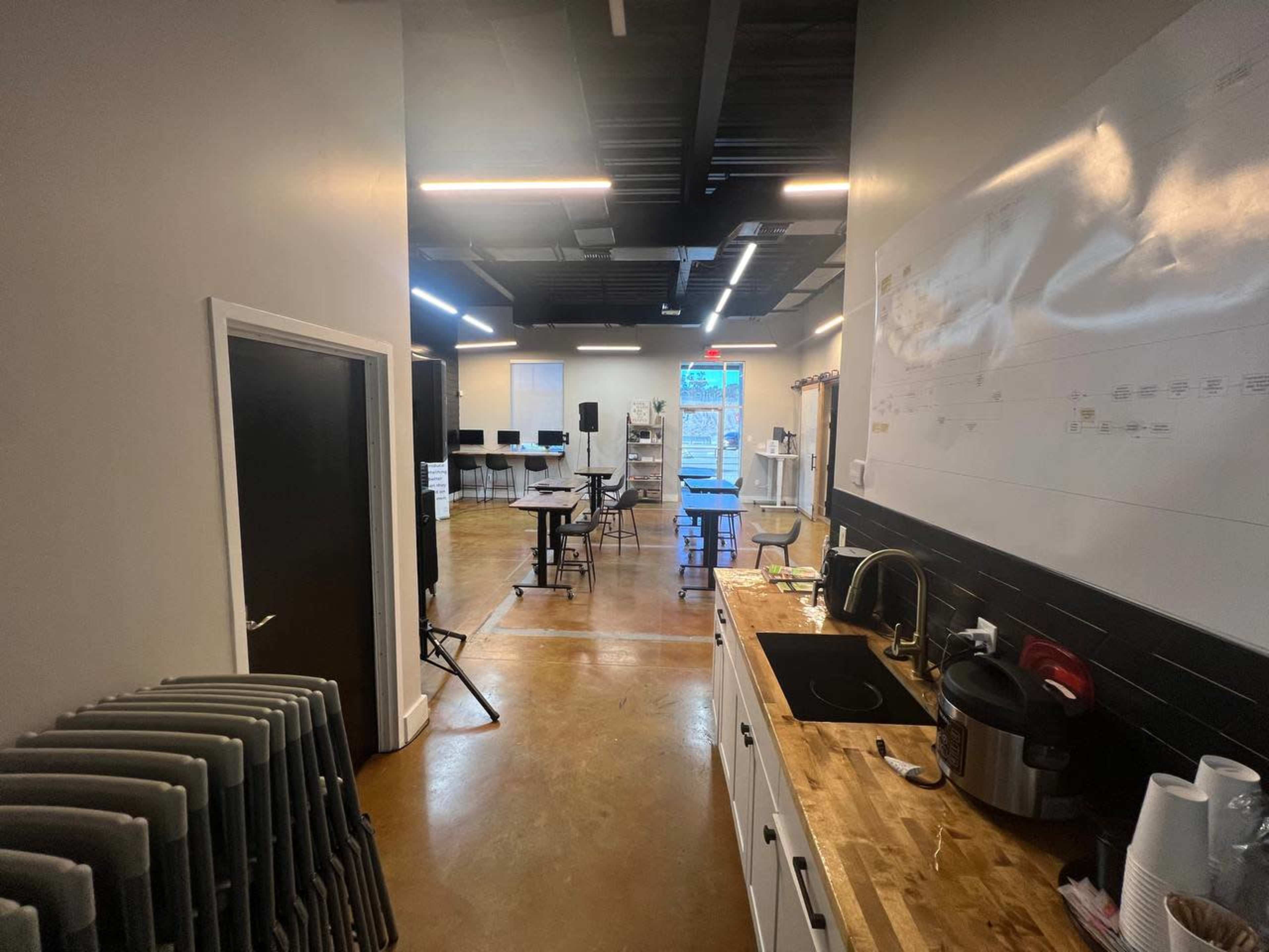 A hallway leading to a large room with tables and chairs, featuring a kitchen area on one side.