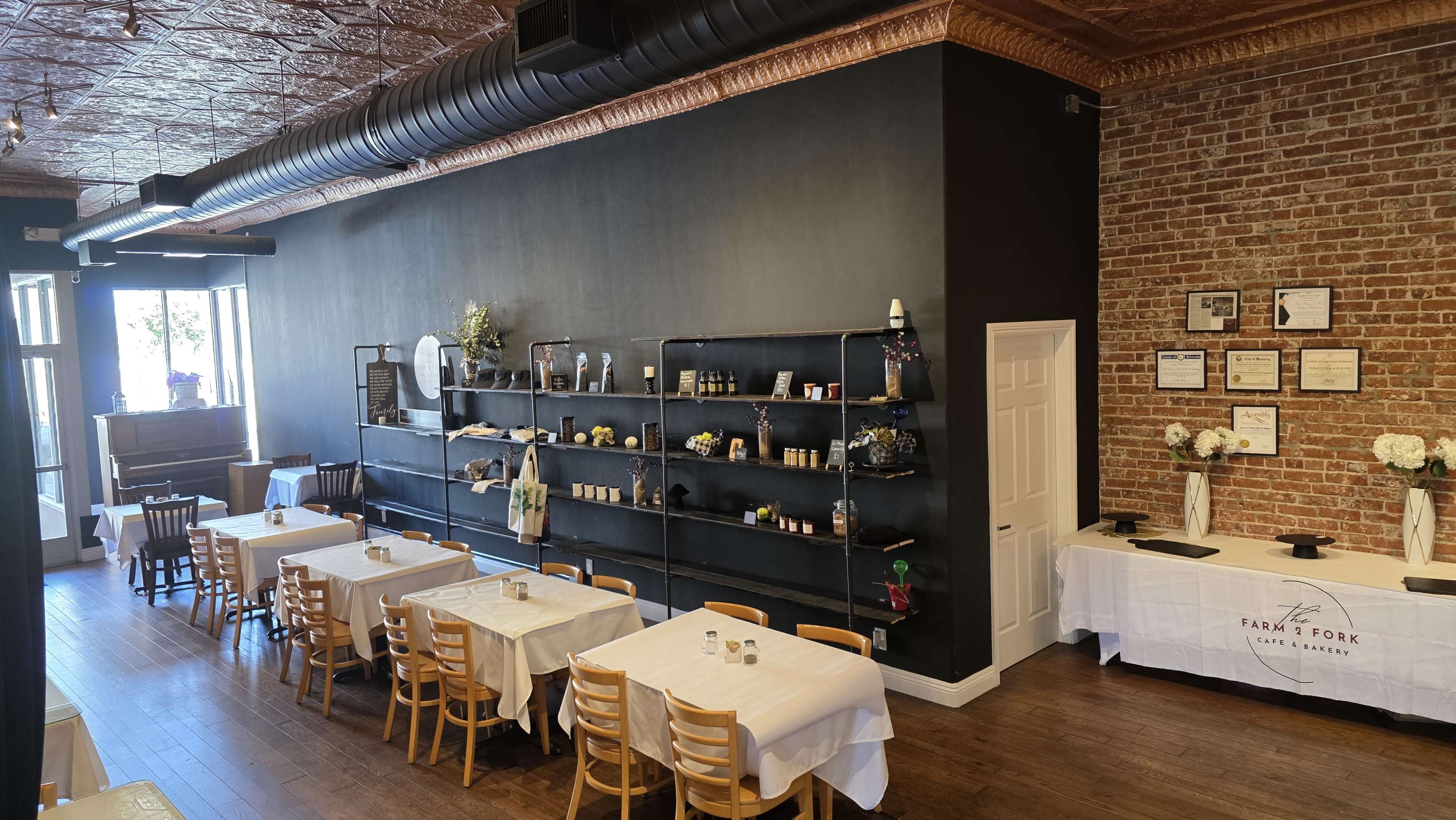 The image shows a restaurant interior with wooden tables covered in white tablecloths, black shelves displaying various items, and a wall featuring exposed brick and decorative elements.