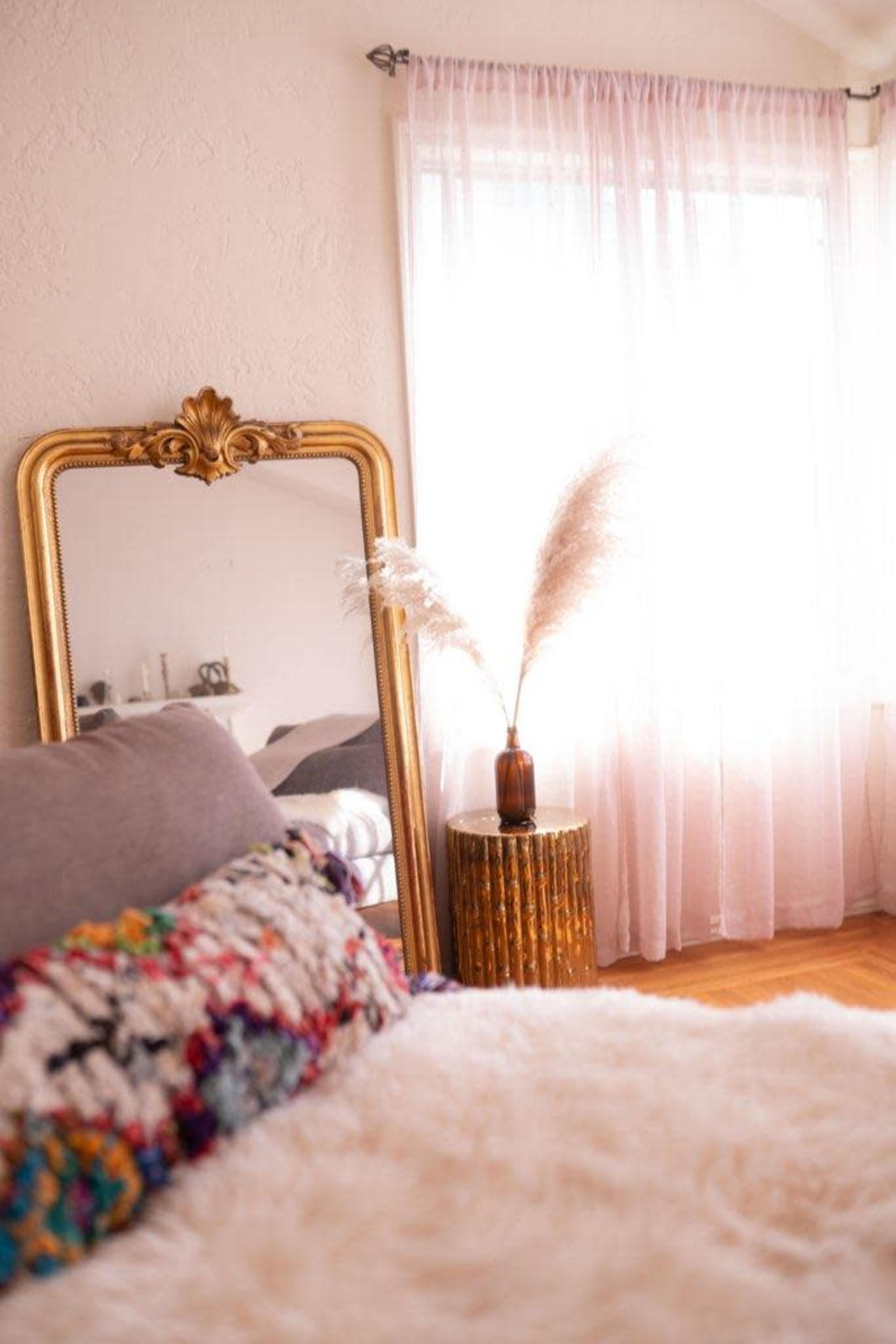 A bedroom interior features a large ornate mirror, a decorative vase with pampas grass on a side table, and sunlight filtering through sheer curtains.