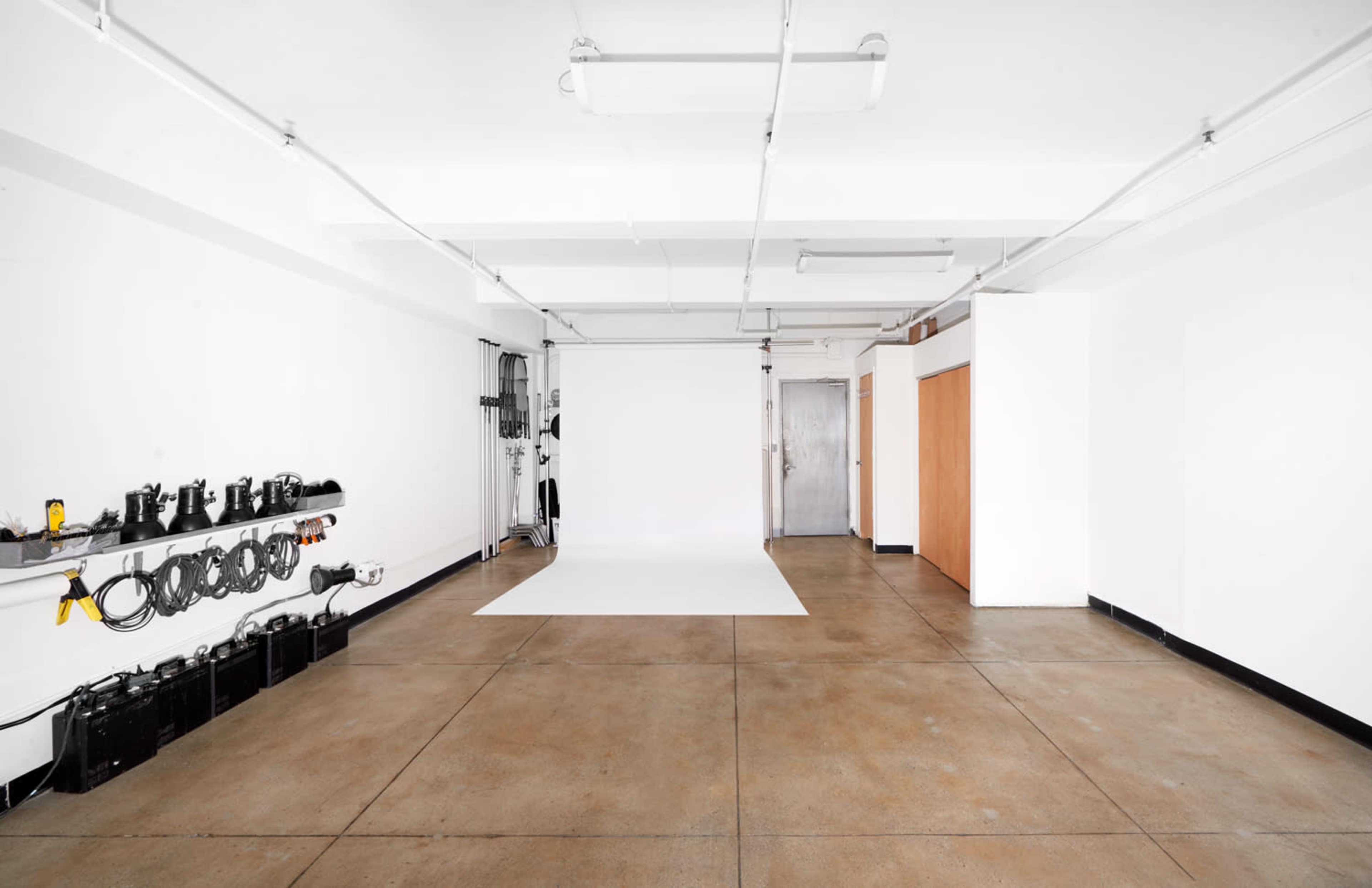 A spacious, empty studio with a white backdrop and industrial flooring, equipped with lighting and equipment mounted along the wall.