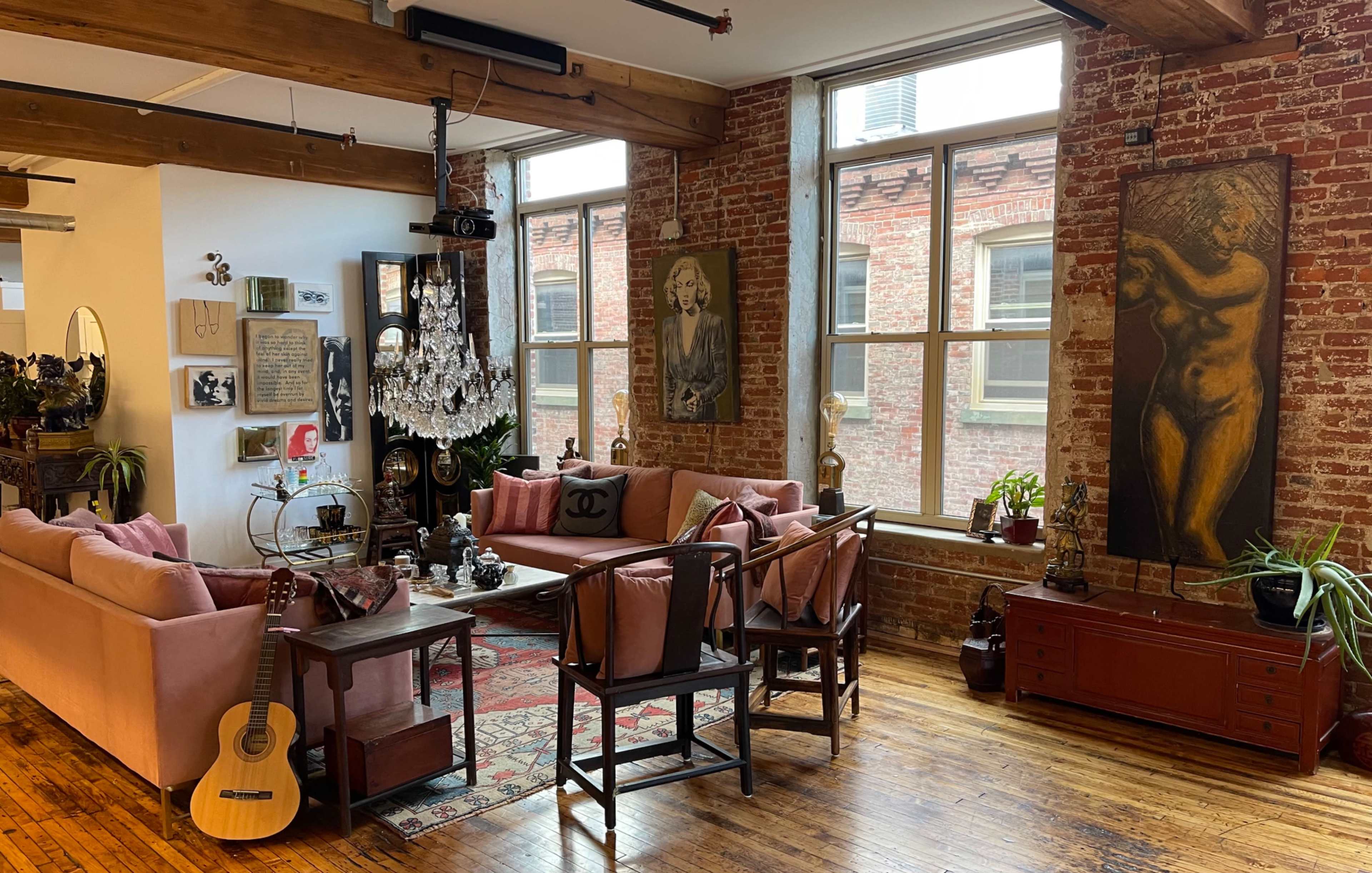 The living space features exposed brick walls, large windows, and a mix of vintage furniture, including pink sofas and a wooden coffee table.