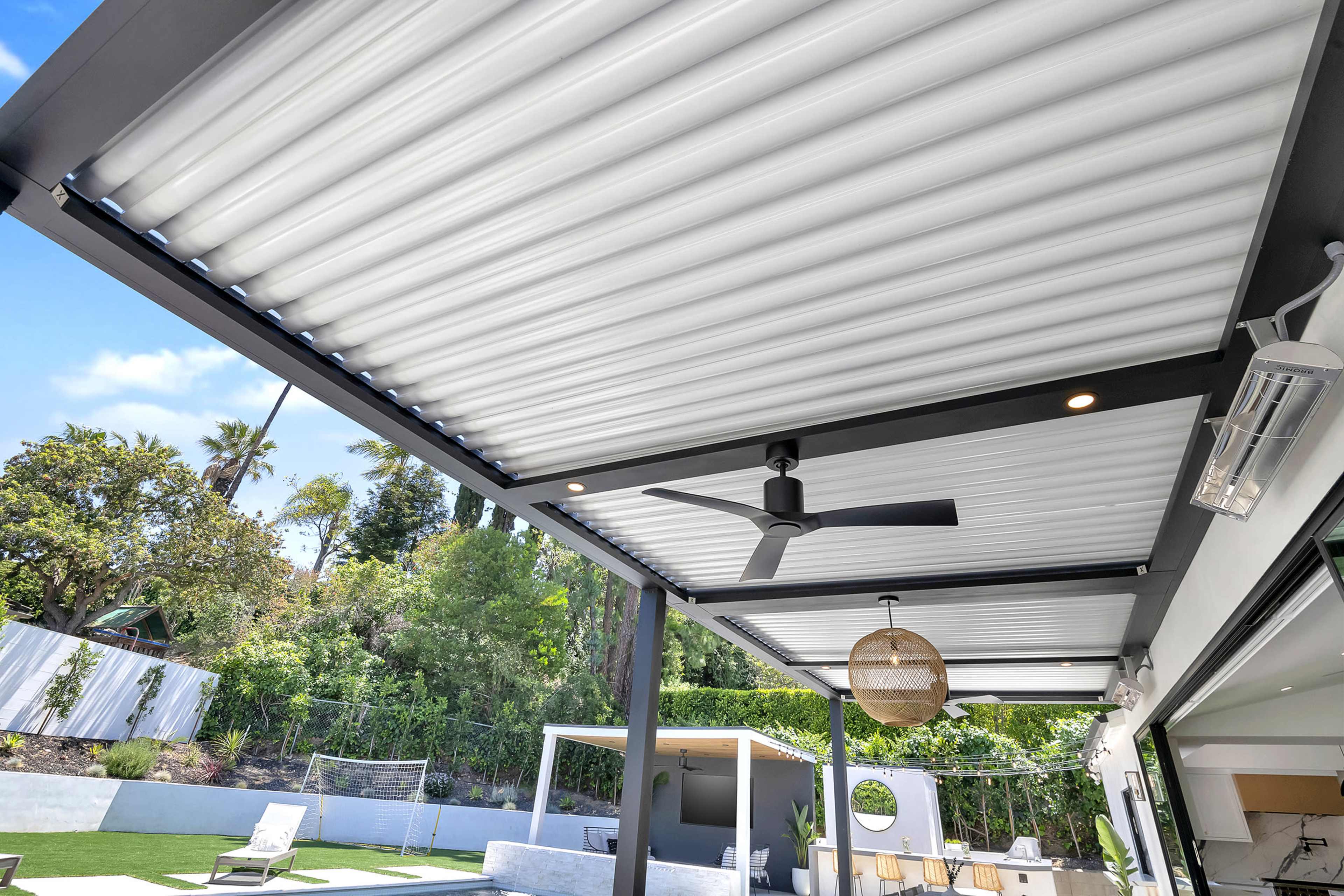 A backyard patio with a retractable roof, featuring a ceiling fan, outdoor lighting, and a view of a landscaped garden and seating area below.