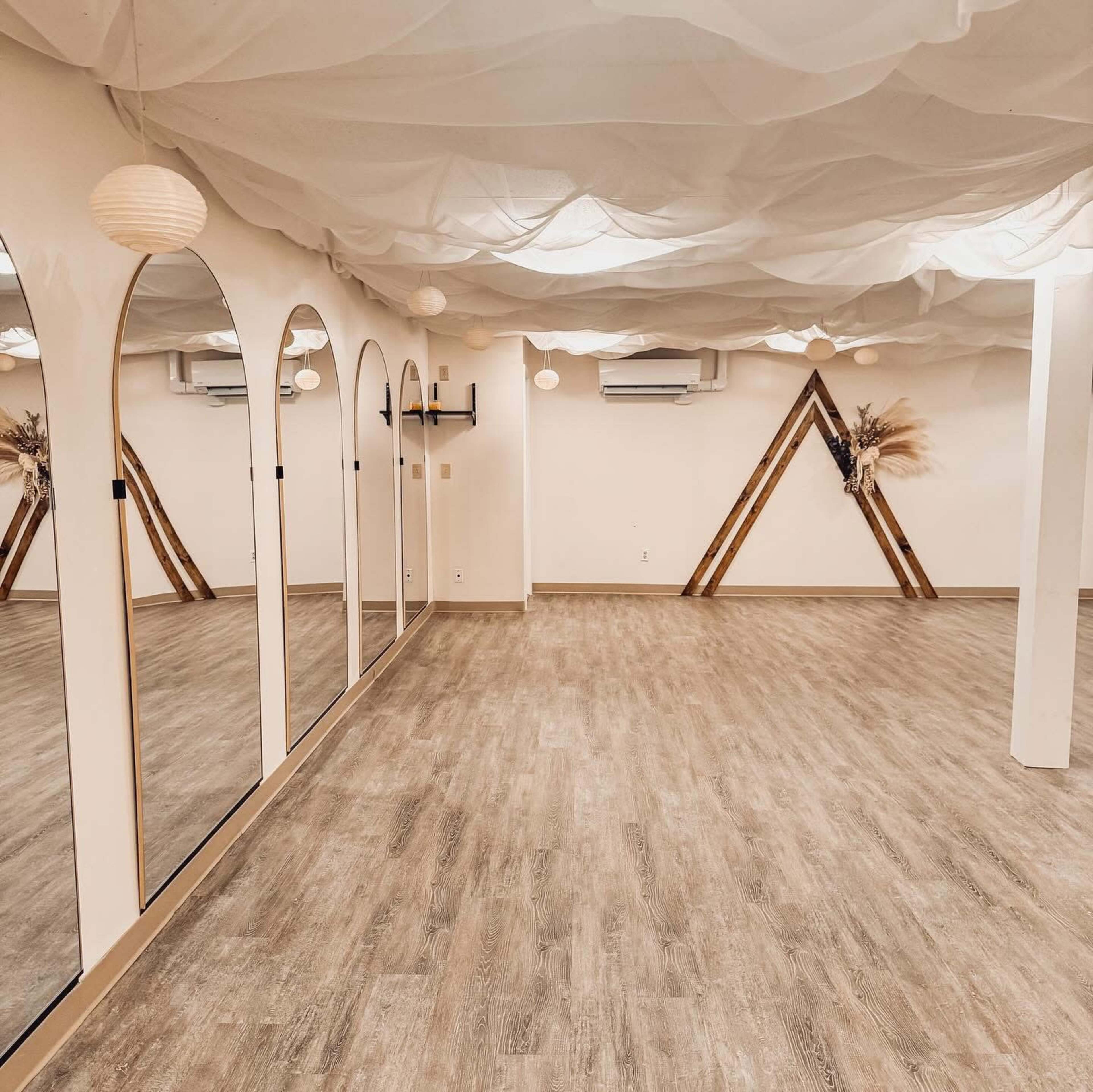 The image shows a spacious room with wooden flooring, mirrors on one wall, and decorative triangular arrangements made of wood and dried plants.