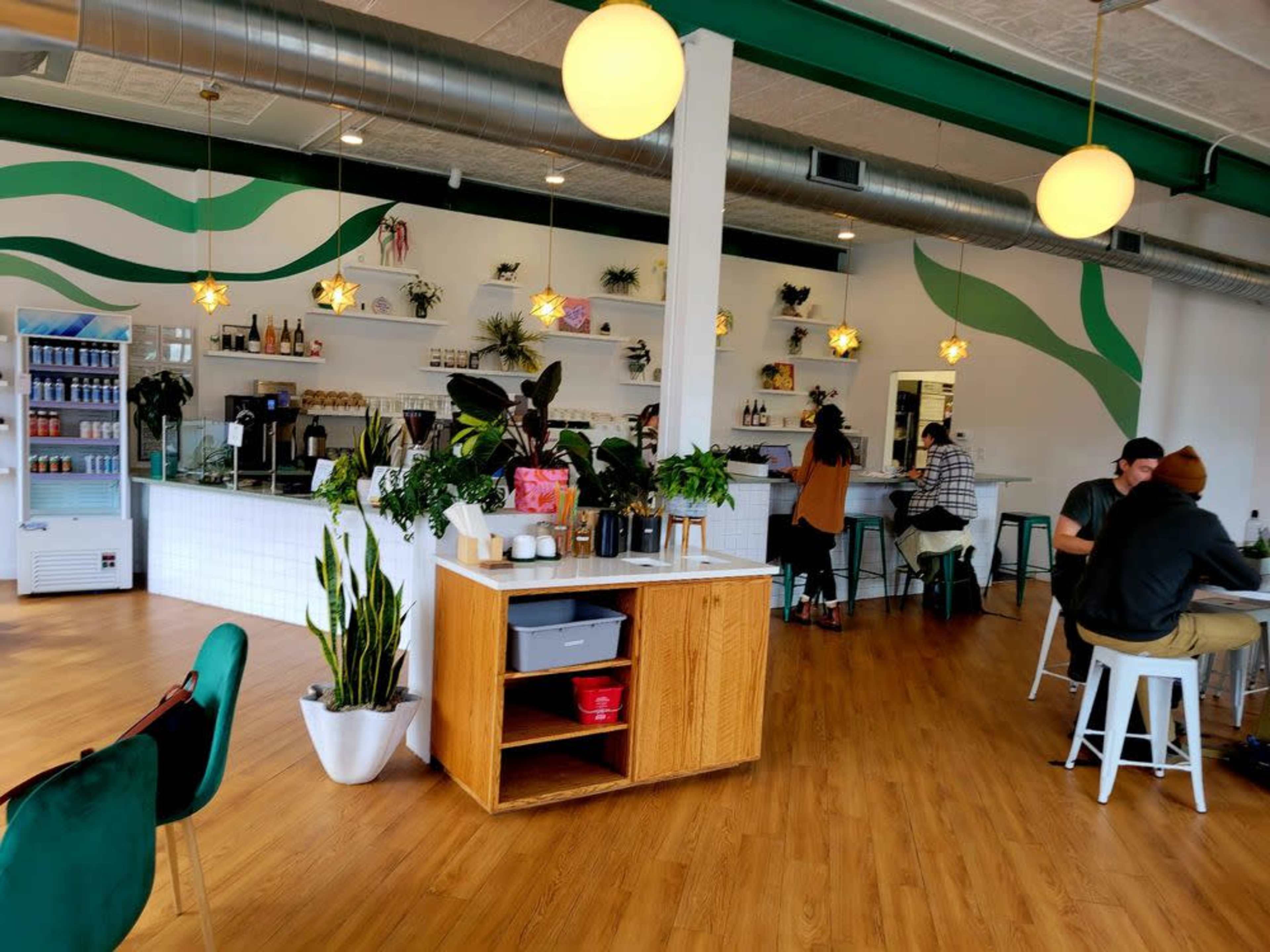 The image shows a modern café interior with a wooden counter, green accents, and potted plants, where customers sit at tables and enjoy their drinks.