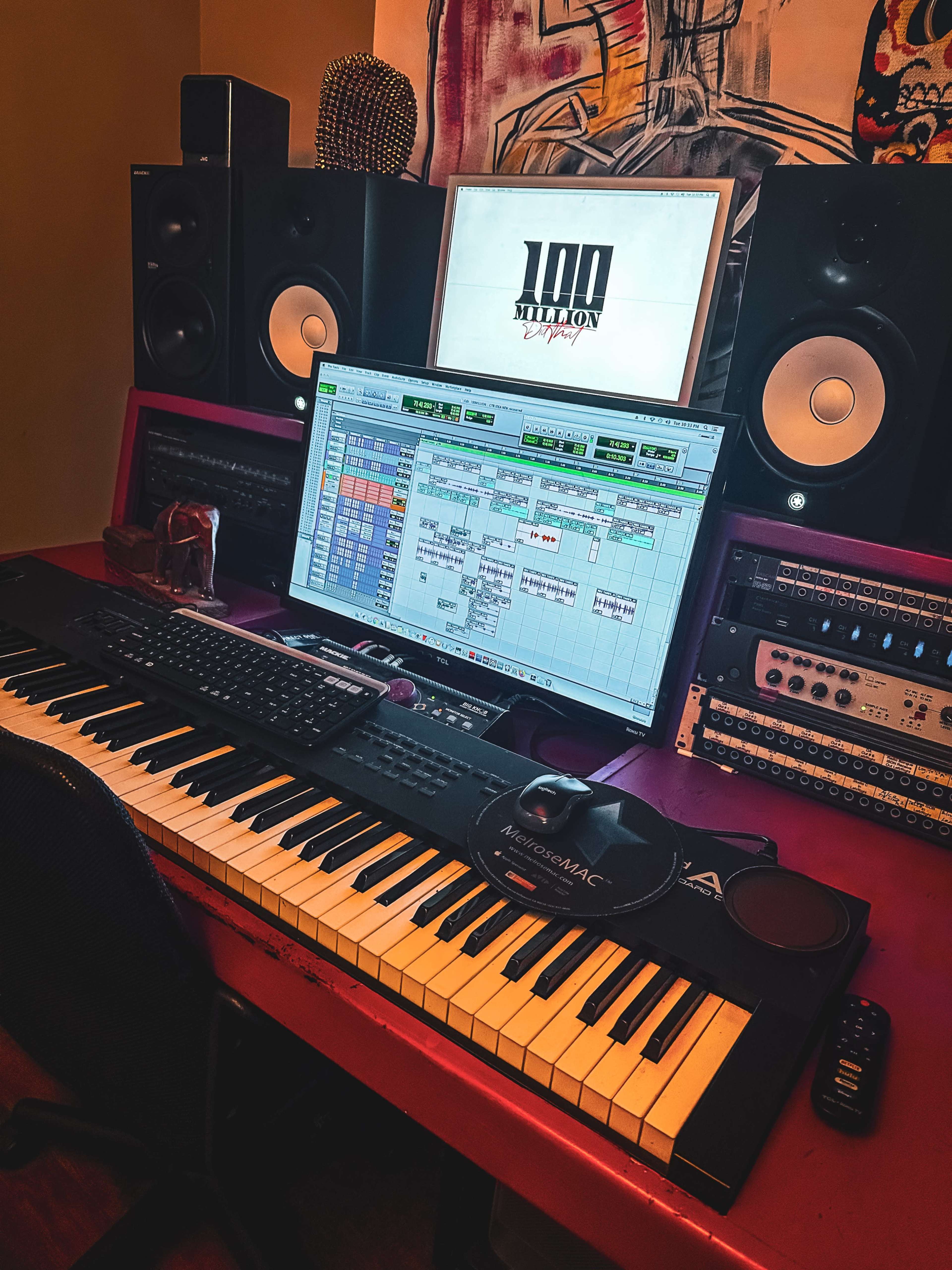 The image shows a music studio setup with a keyboard, computer monitor displaying a music project, and large speakers on either side of the monitor.