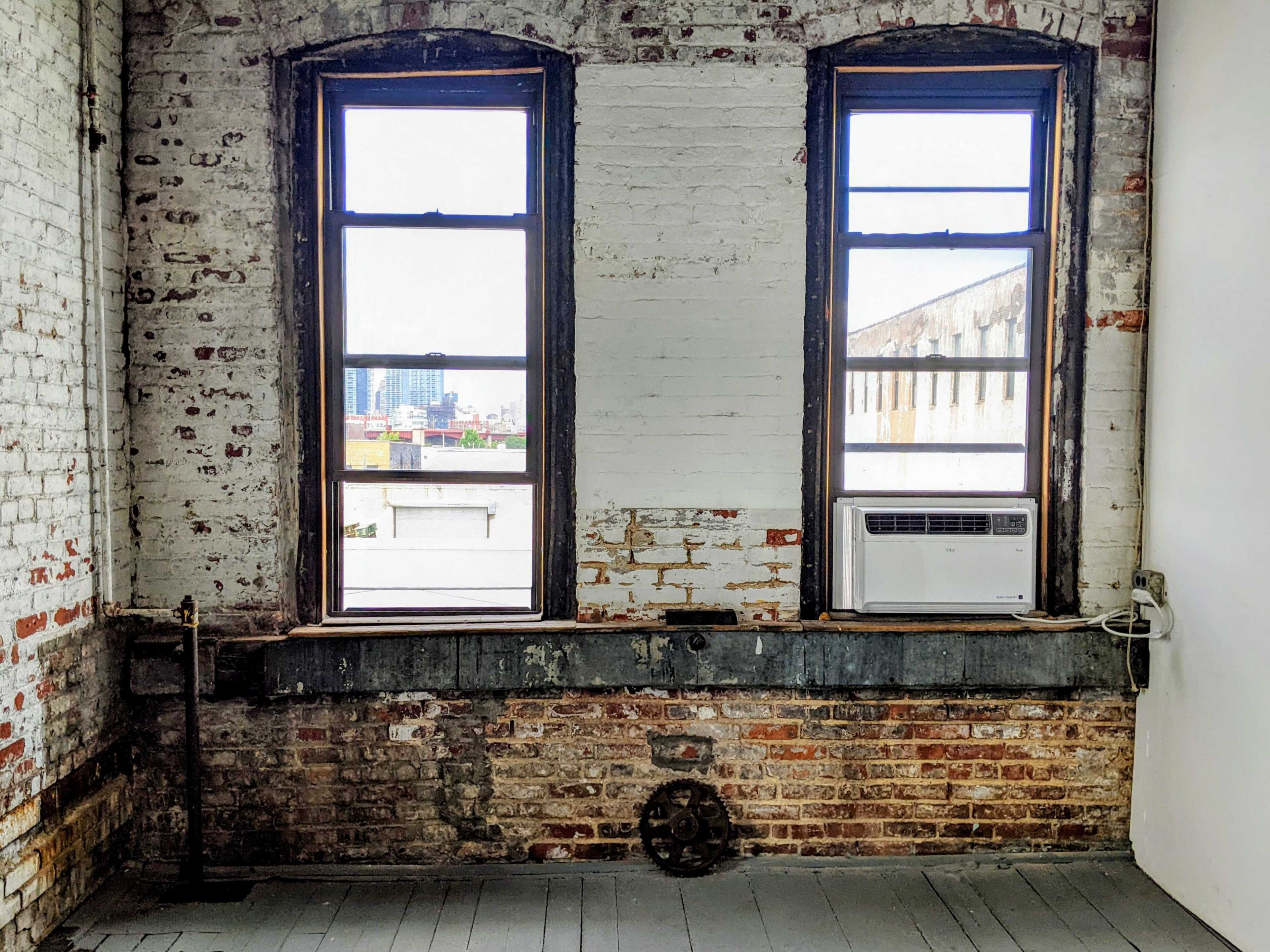 A room with two tall windows, exposed brick walls, and an air conditioning unit placed beneath one of the windows.