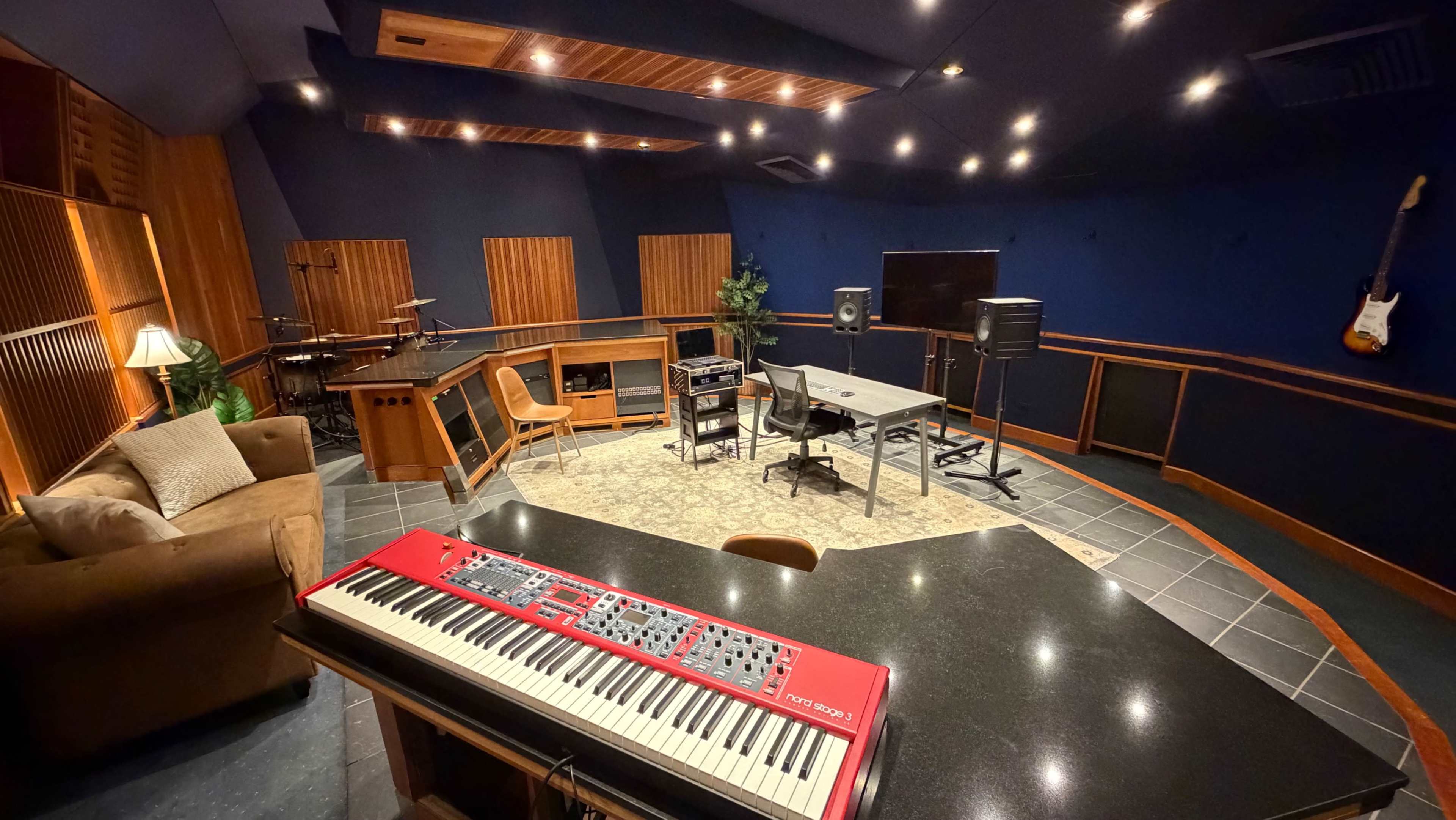 A music recording studio features a red keyboard on a countertop, surrounded by instruments, desks, and studio equipment in a spacious room with acoustic panels.