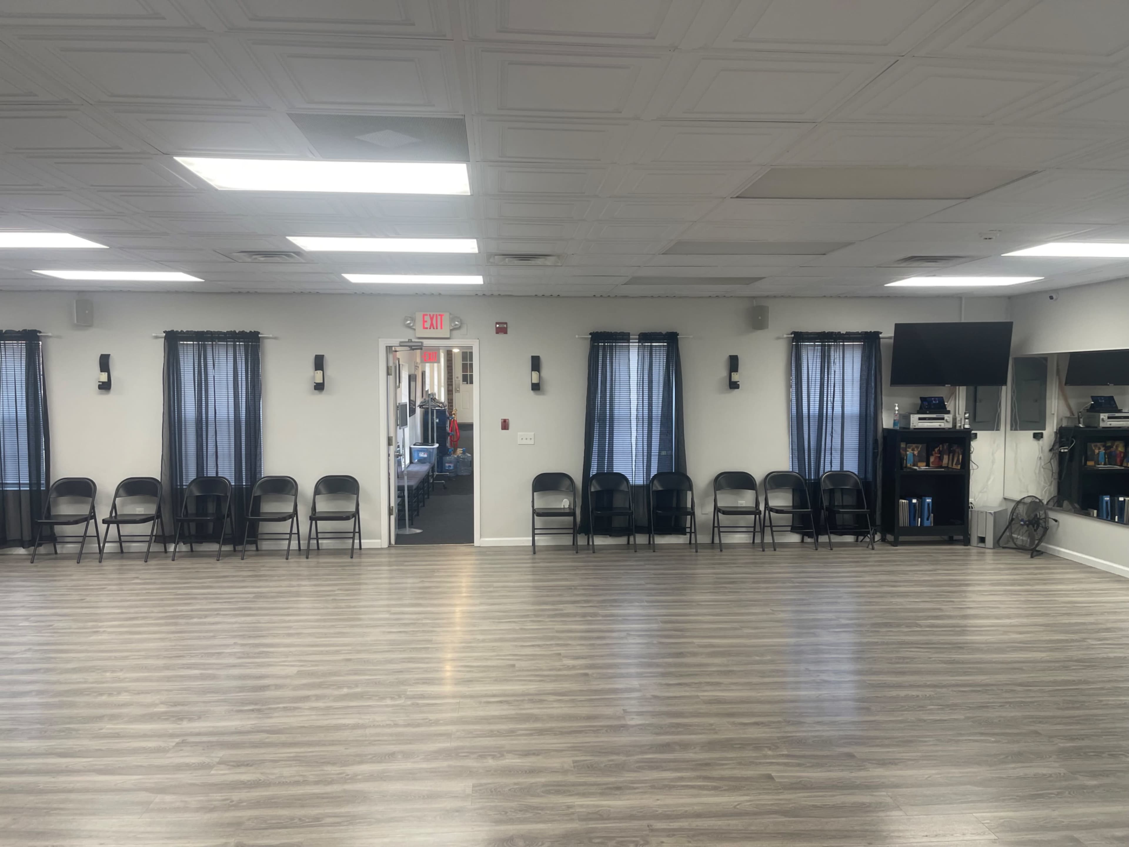 A spacious room with multiple chairs arranged in a line along one wall, large windows with dark curtains, and a doorway leading to another area.