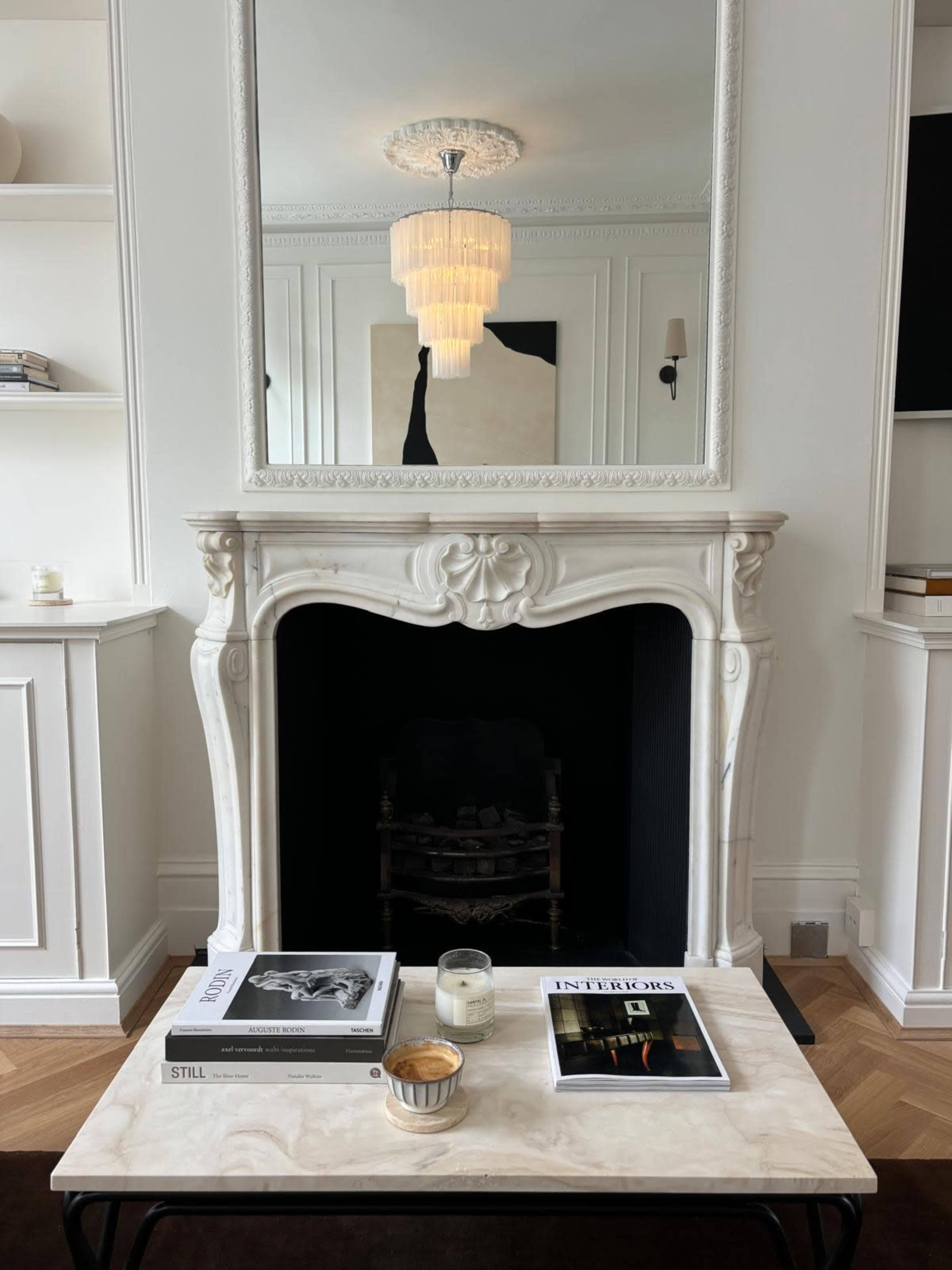 The image shows a minimalist living room with a stylish marble fireplace, a mirror above it, and a coffee table adorned with books and a candle.