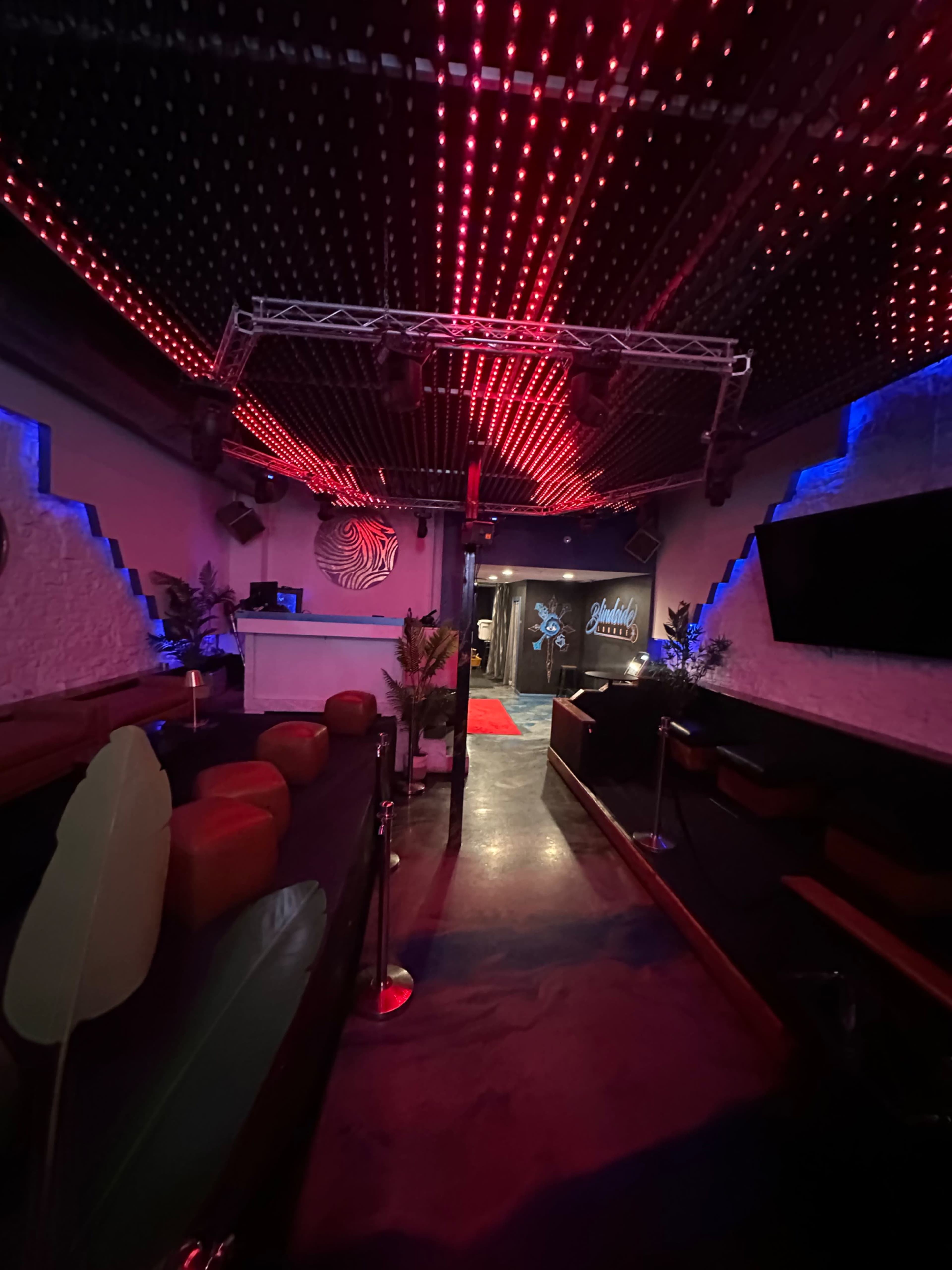 The image shows a dimly lit bar or lounge featuring red and blue LED lights, a small stage area, and a bar counter at the back.