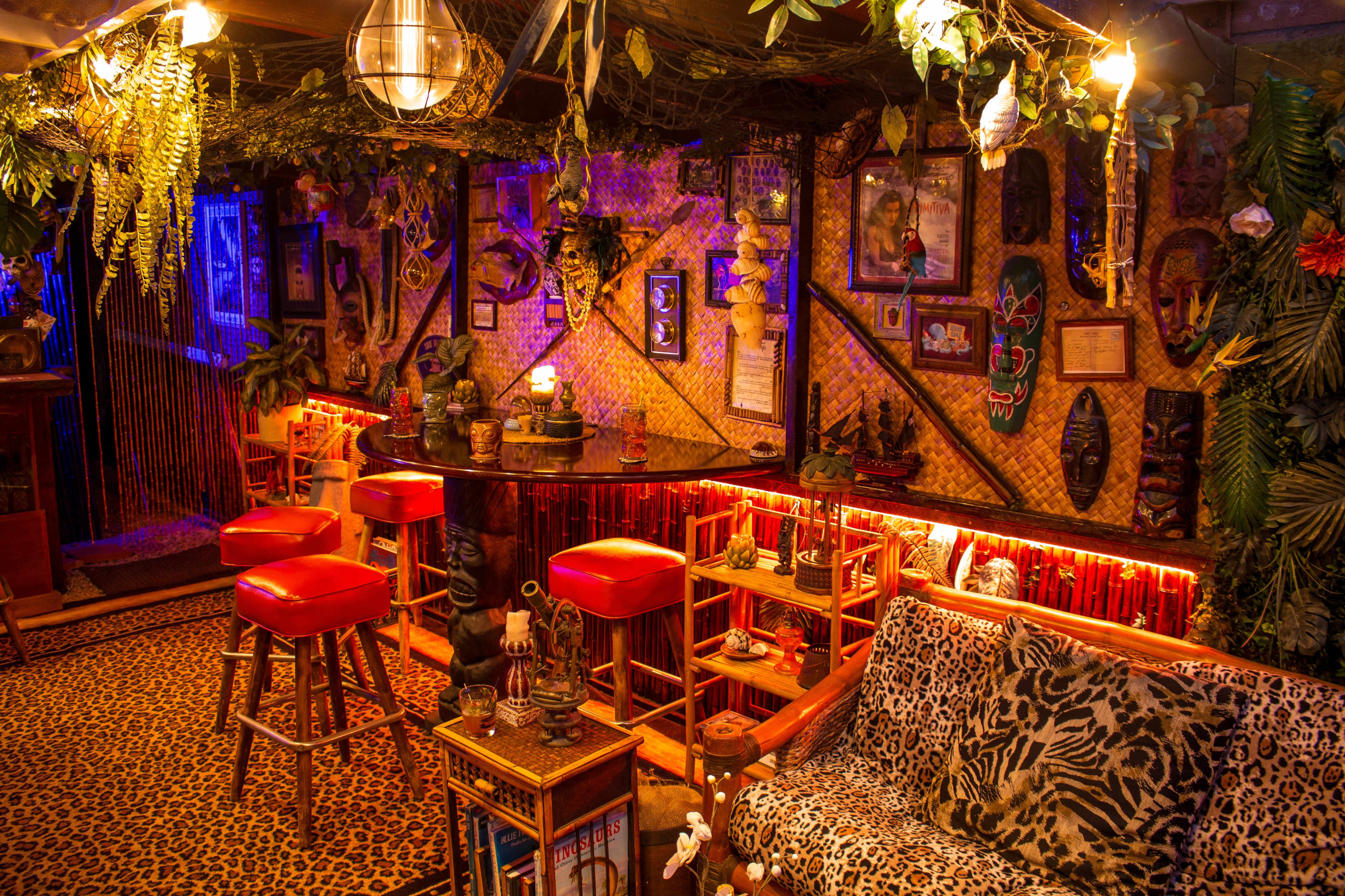 A decorated indoor lounge with a bar, surrounded by tropical plants, animal prints, and various framed artworks.