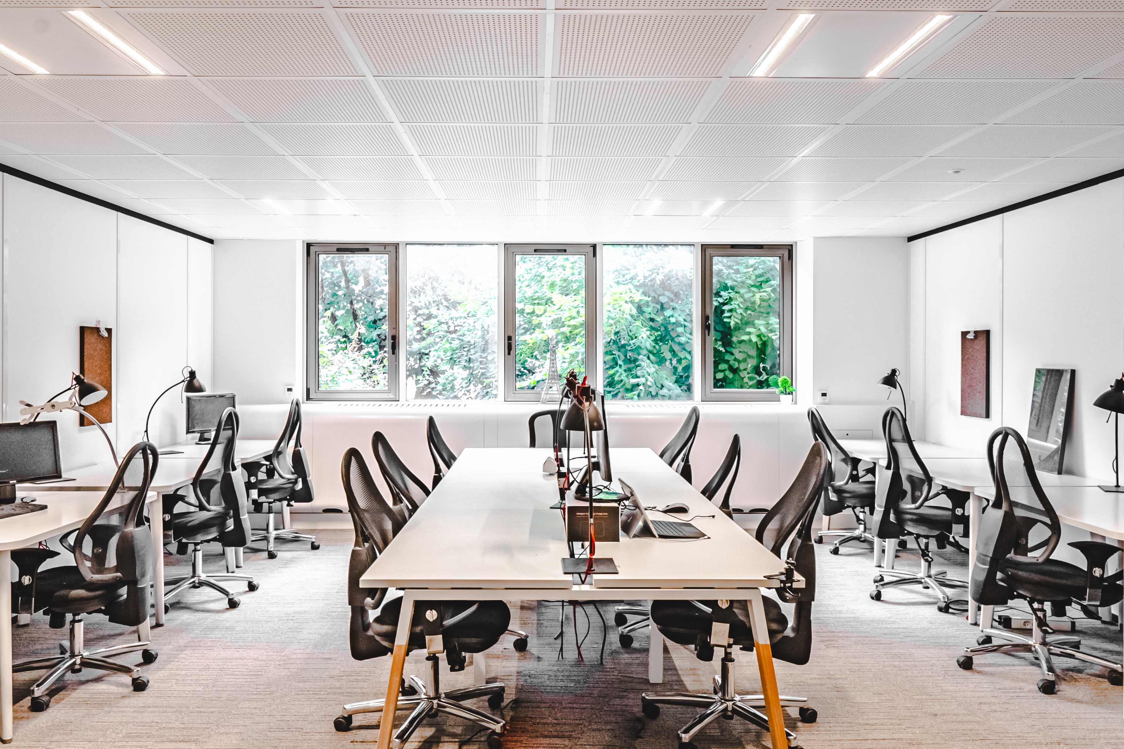 The image shows a modern office space with several desks and chairs arranged neatly, and large windows overlooking greenery.