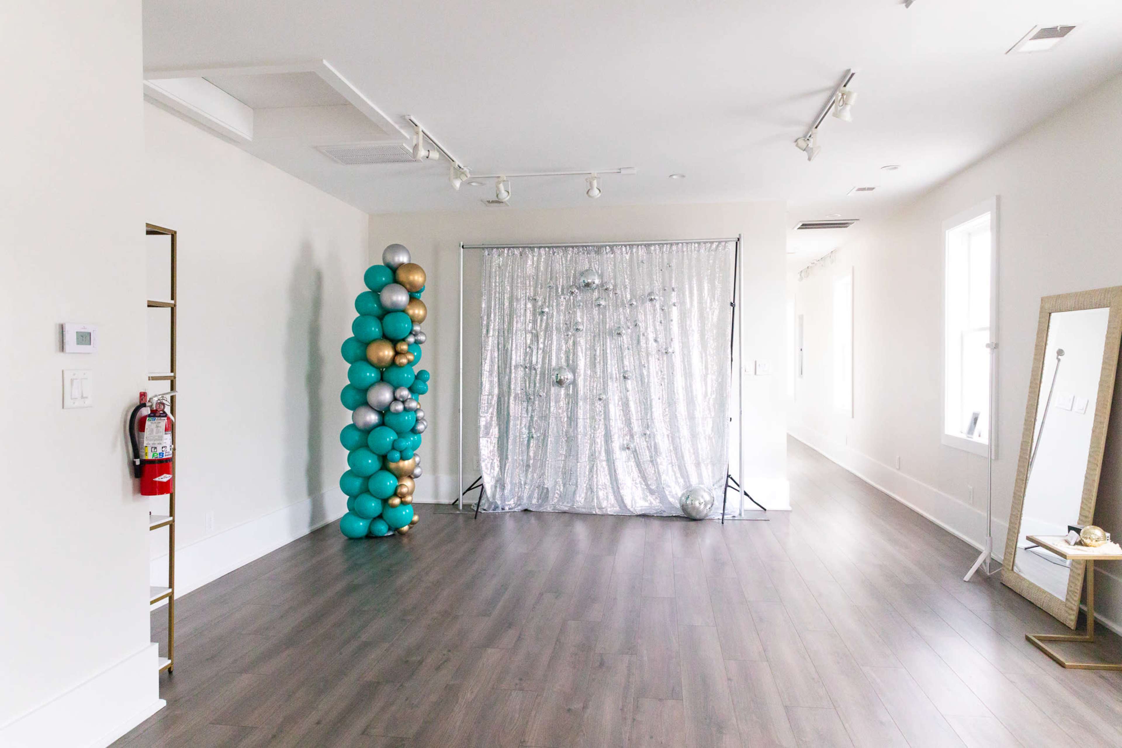 A spacious, well-lit room features a silver backdrop with decorative balloons in teal, gold, and silver arranged to one side.