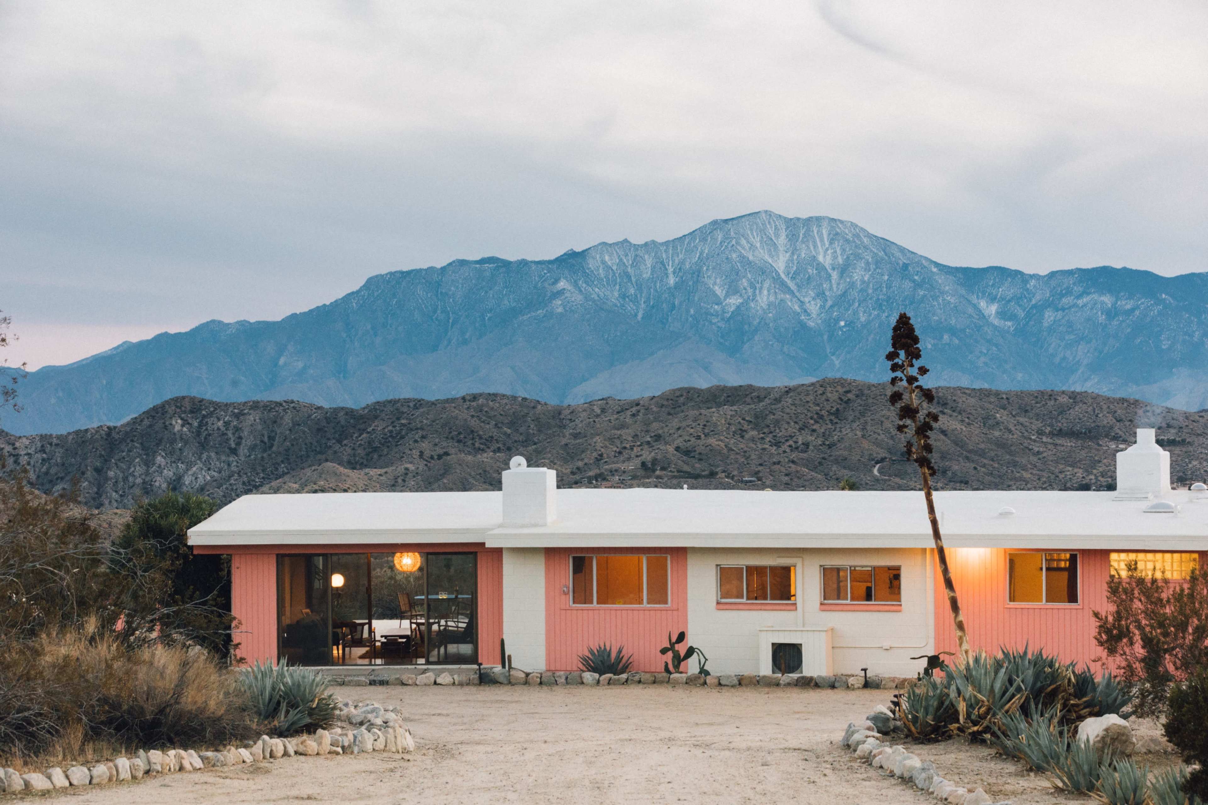 A pink mid-century modern house stands in front of a mountainous landscape with snow-capped peaks.