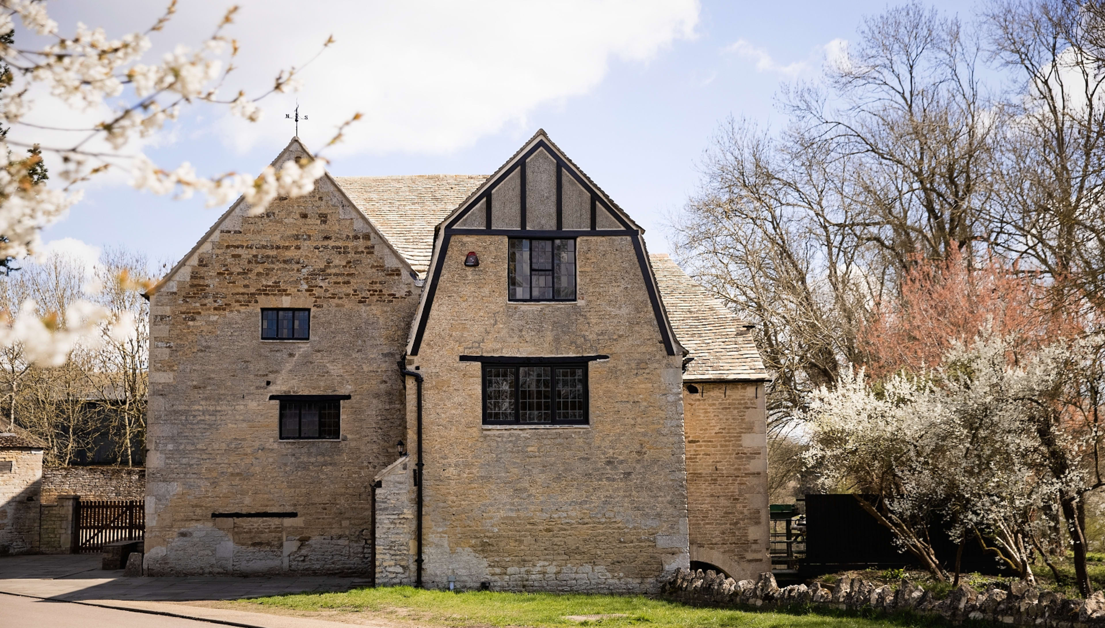 A stone and timber-framed house stands beside a road, surrounded by trees and blooming plants.