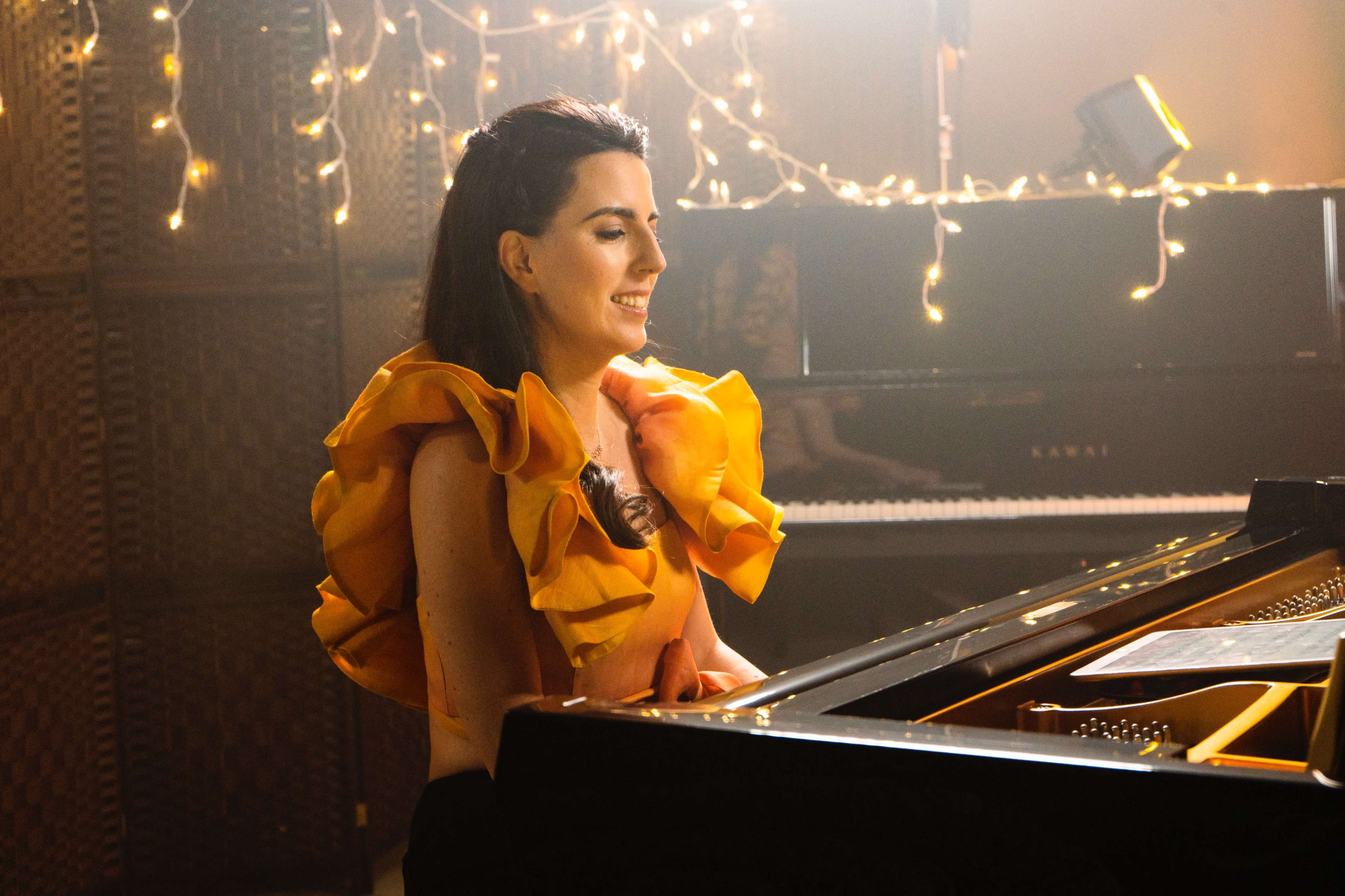 A woman in a yellow ruffled top sits at a grand piano, illuminated by soft lighting and surrounded by twinkling string lights.