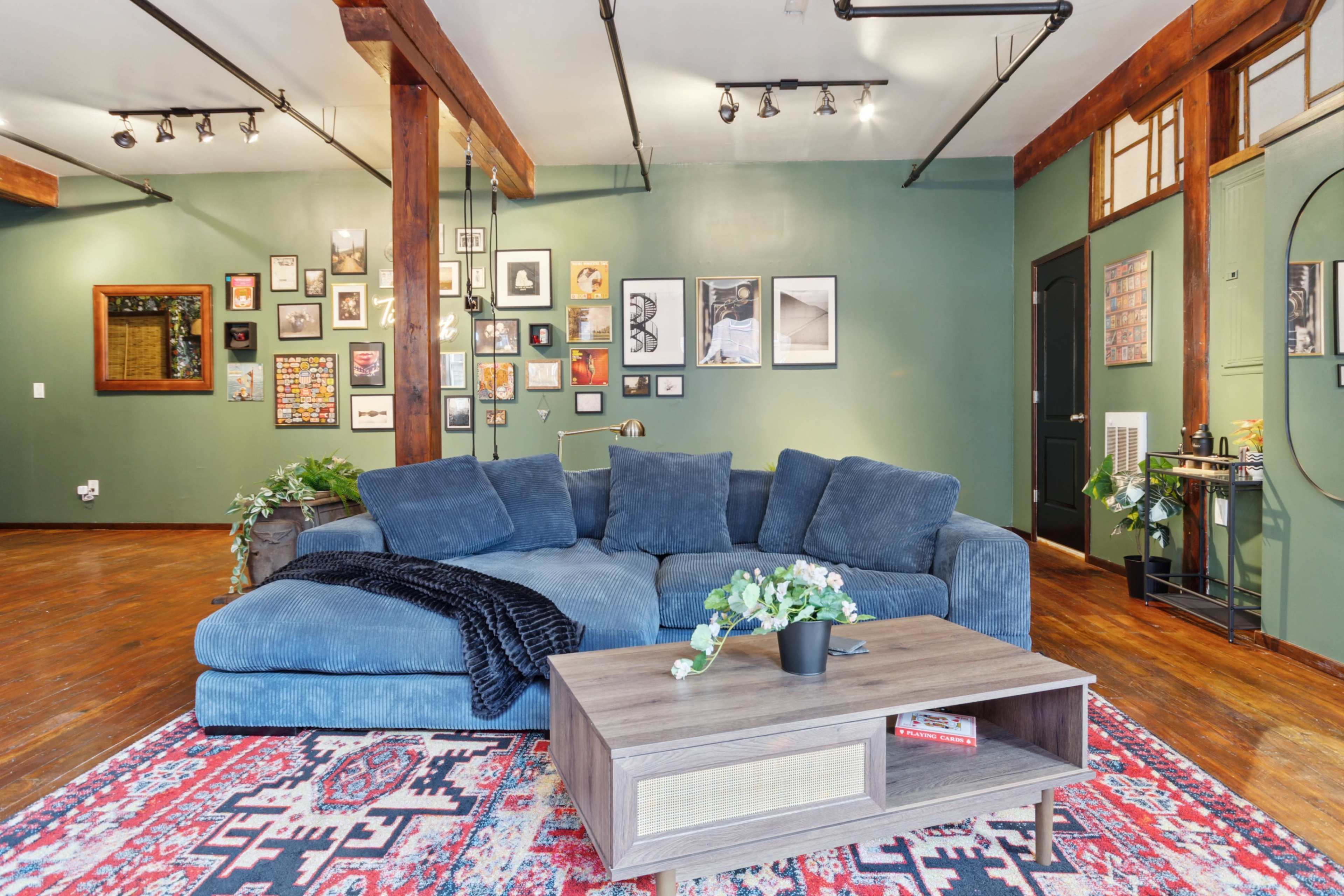 A modern living room features a large, blue sectional sofa, a wooden coffee table adorned with plants, and a gallery wall of framed art against a green backdrop.