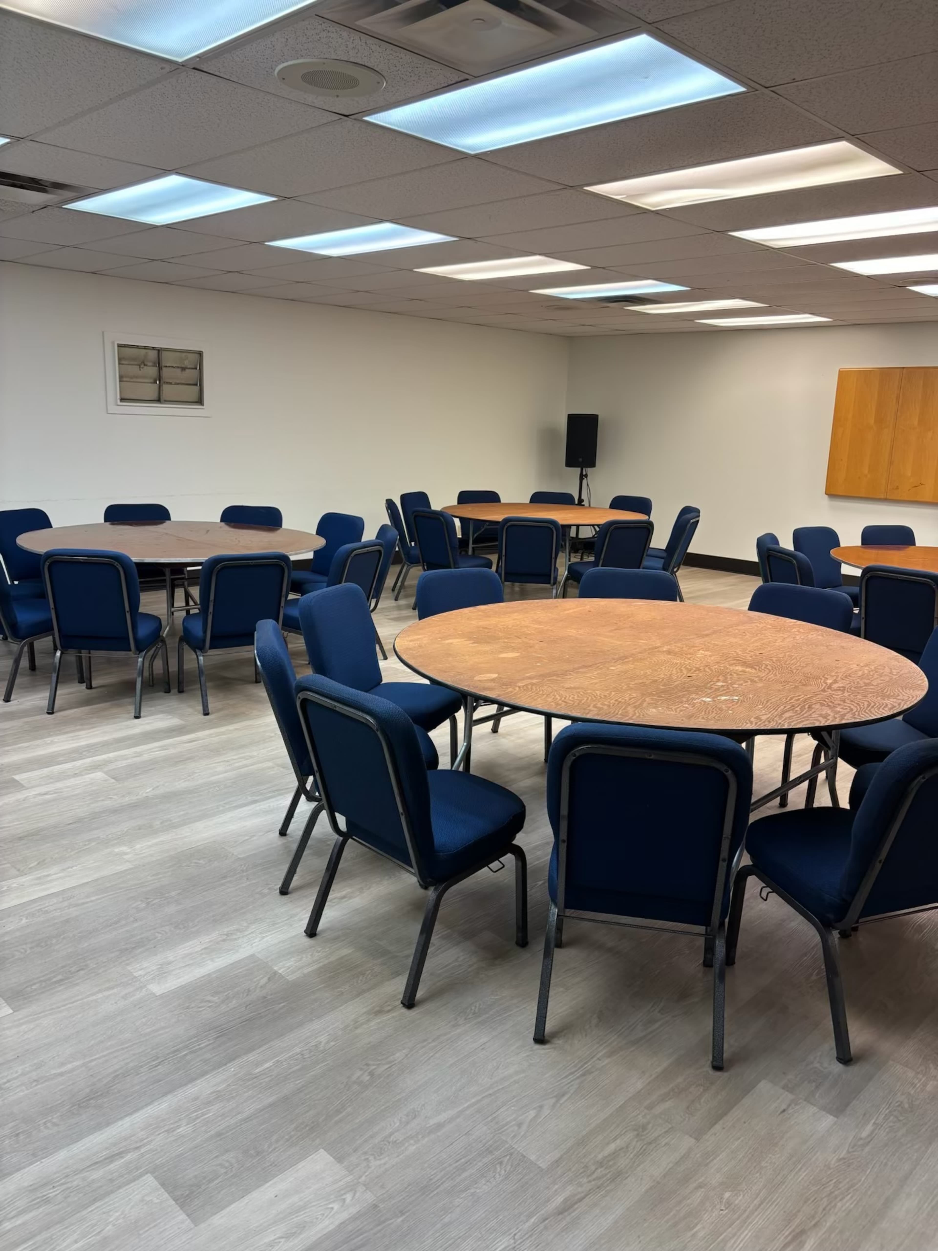 The image shows a meeting room with several round tables surrounded by blue chairs on a wooden floor.