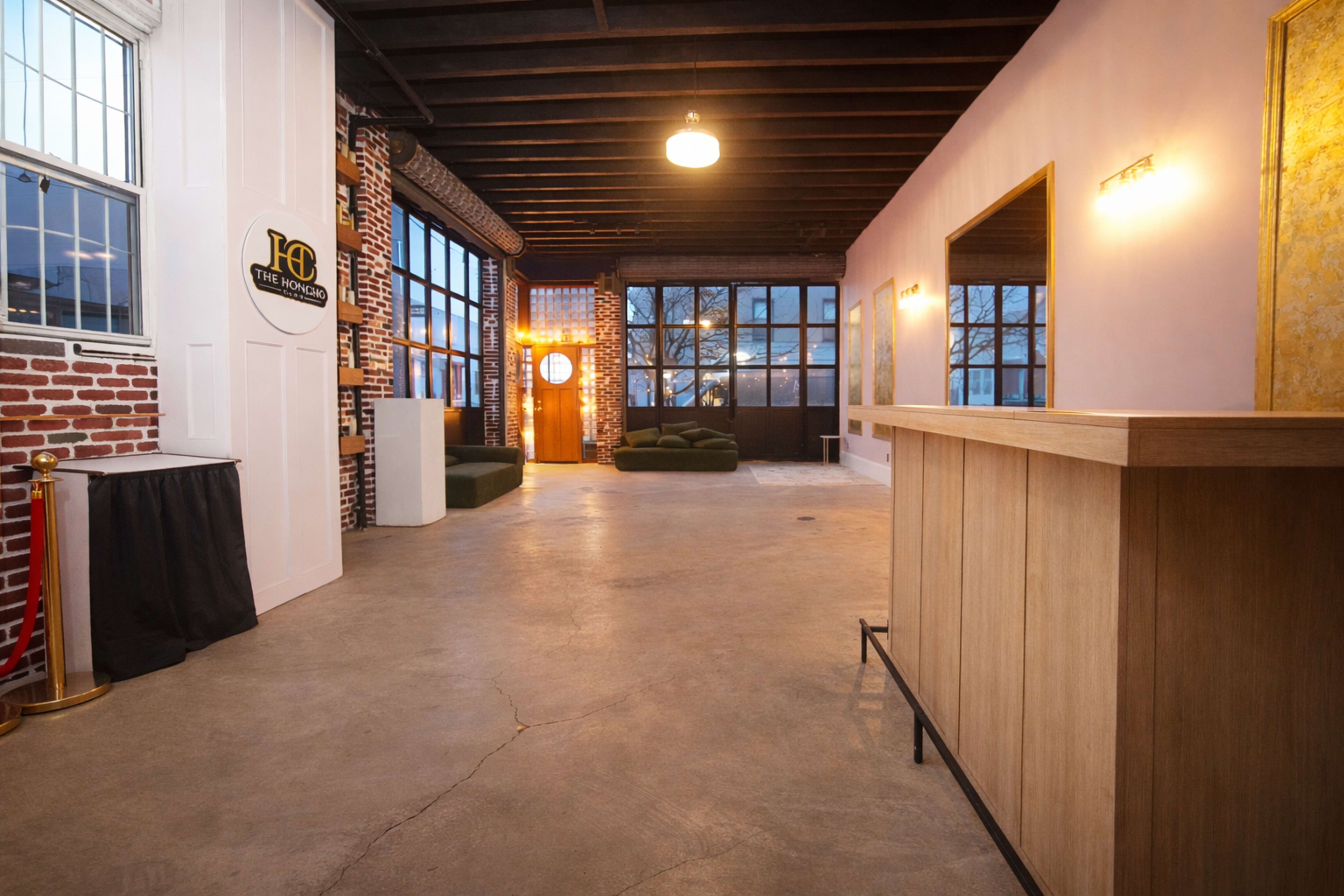 The image shows a spacious, modern interior with exposed brick walls, large windows, and a wooden reception desk.