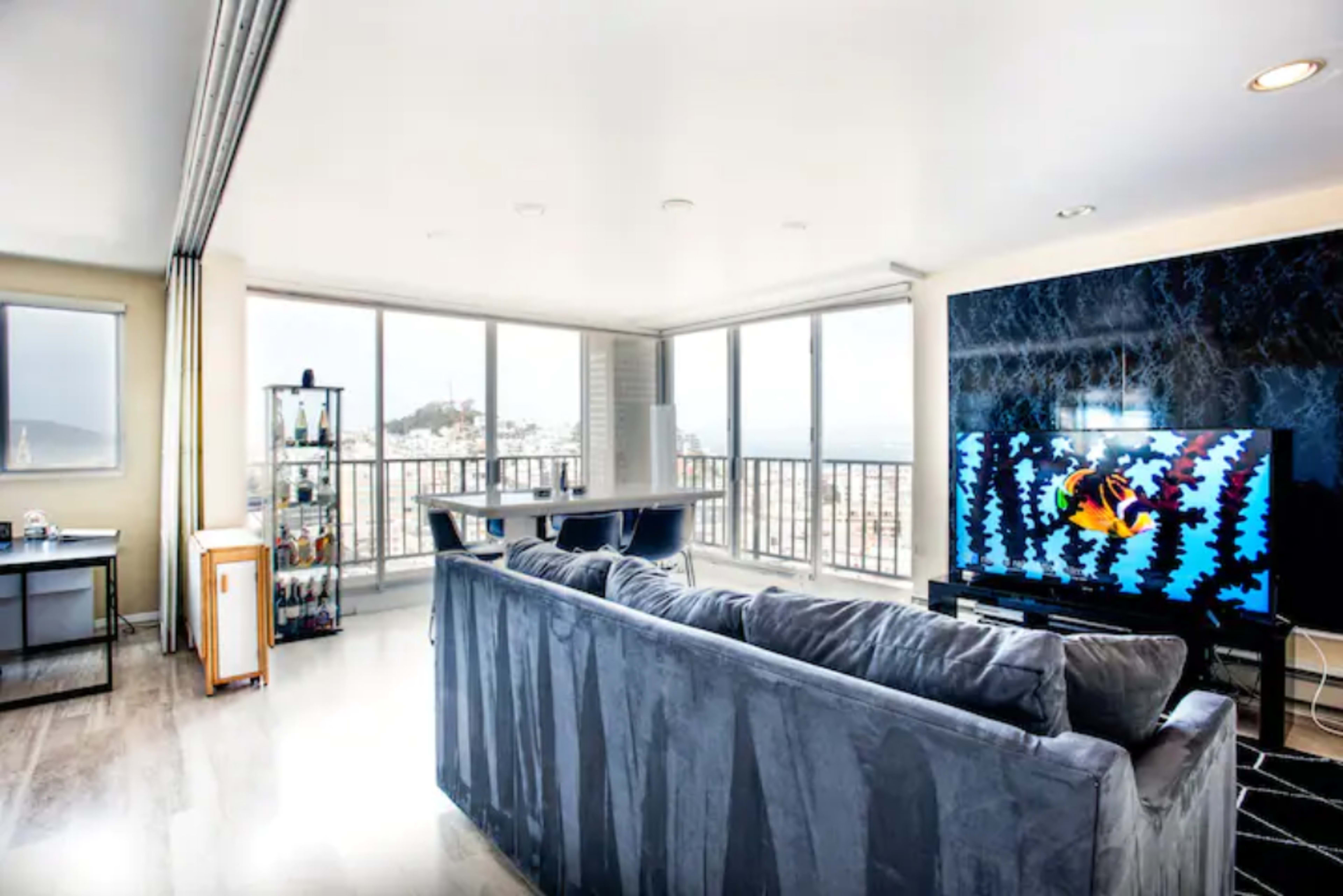 A modern living room with a large couch facing a wall-mounted television and large windows offering a view of the city.