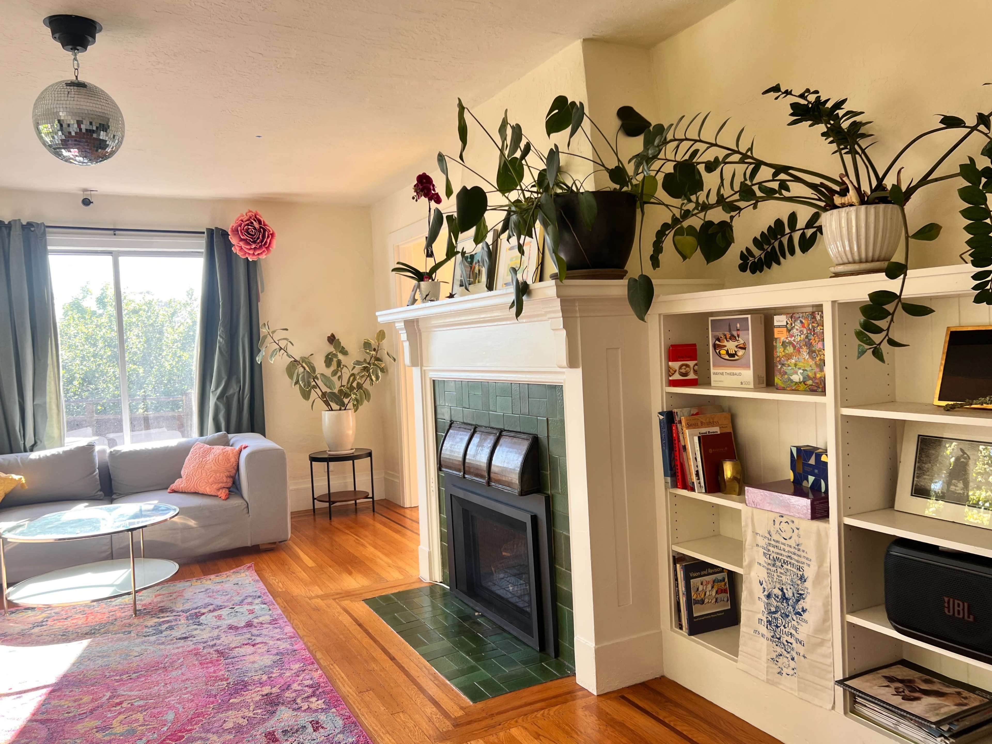The image shows a cozy living room featuring a gray couch, a circular glass coffee table, a fireplace with green tiles, and a bookshelf with colorful books and decorations.