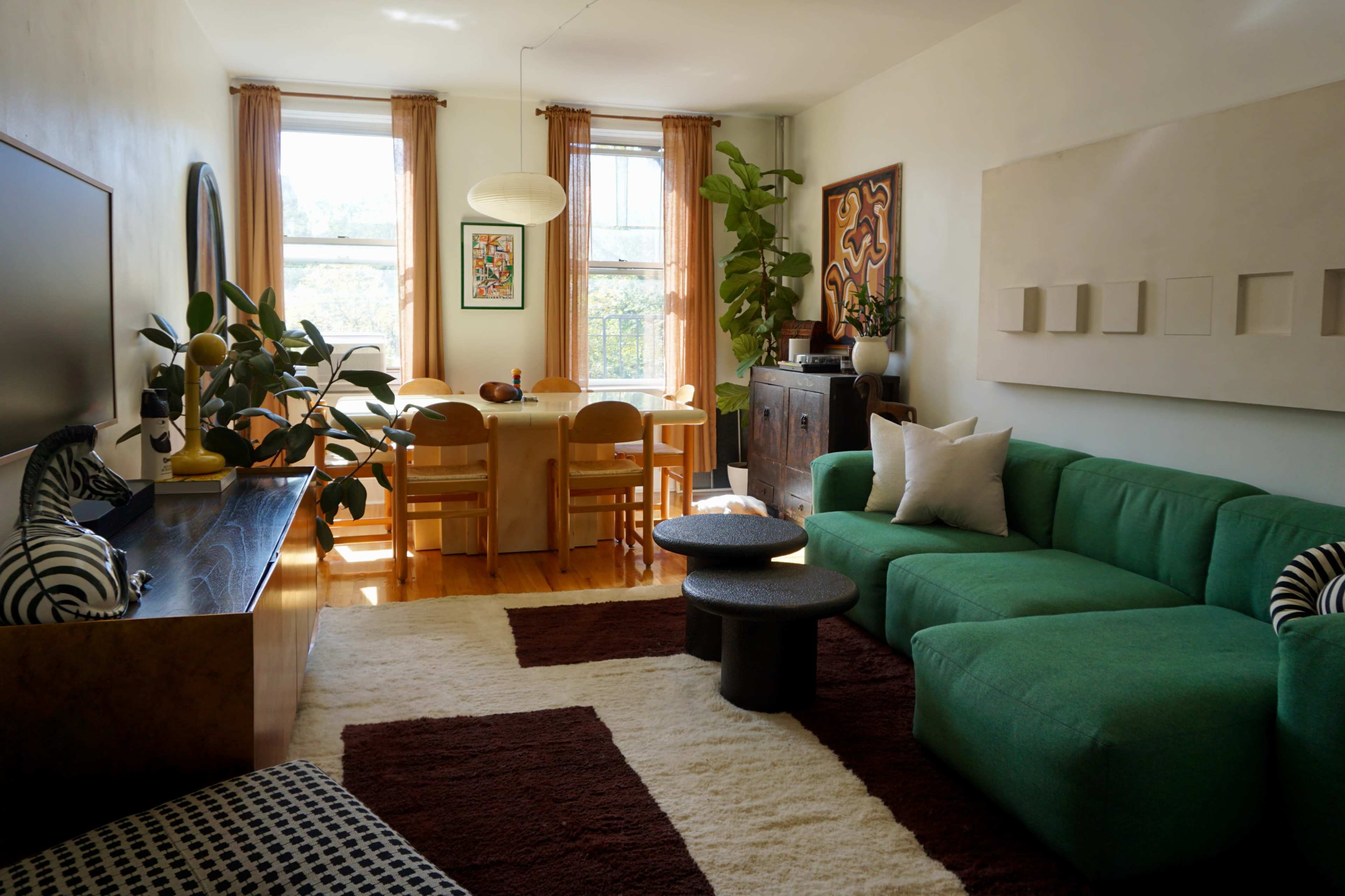 The room features a living area with a green sectional sofa, a black and white coffee table, and a dining table with wooden chairs, accompanied by large windows and plants.