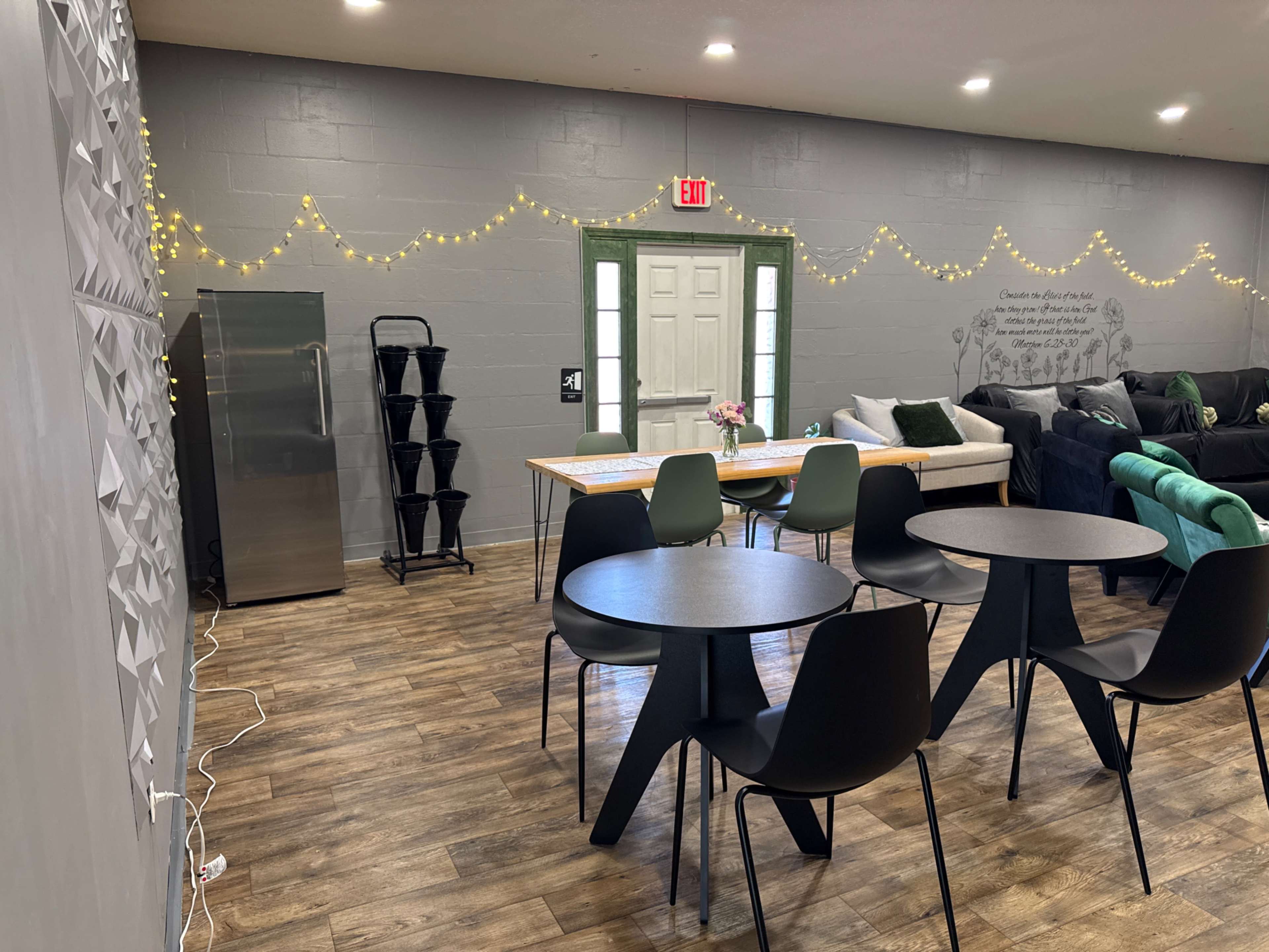 The image shows a cozy communal space with two black tables, green and black chairs, a refrigerator, and decorative string lights on the walls.