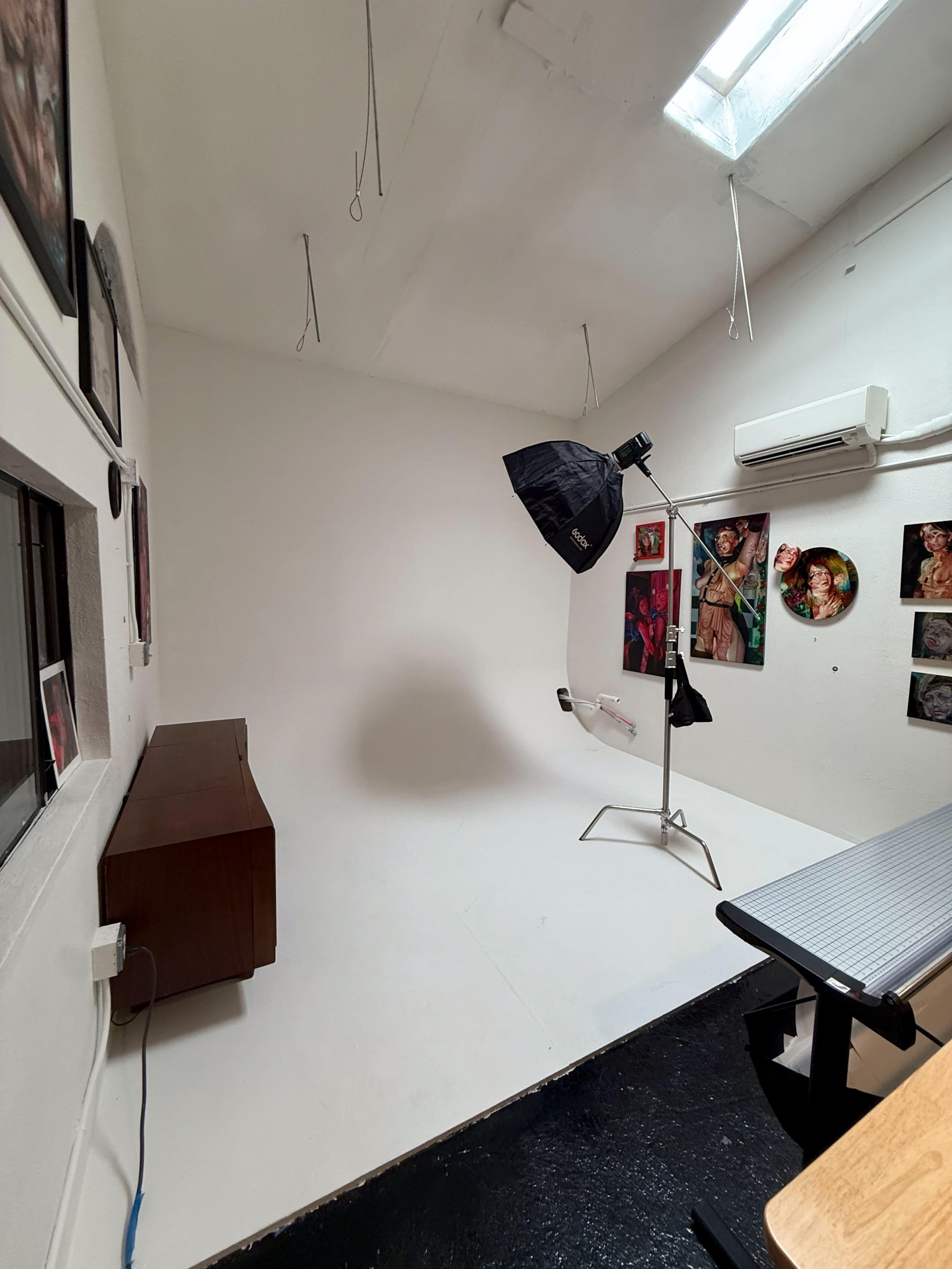 The image shows a brightly lit, minimalist photography studio with a large softbox light, a white backdrop, and several framed photographs on the walls.