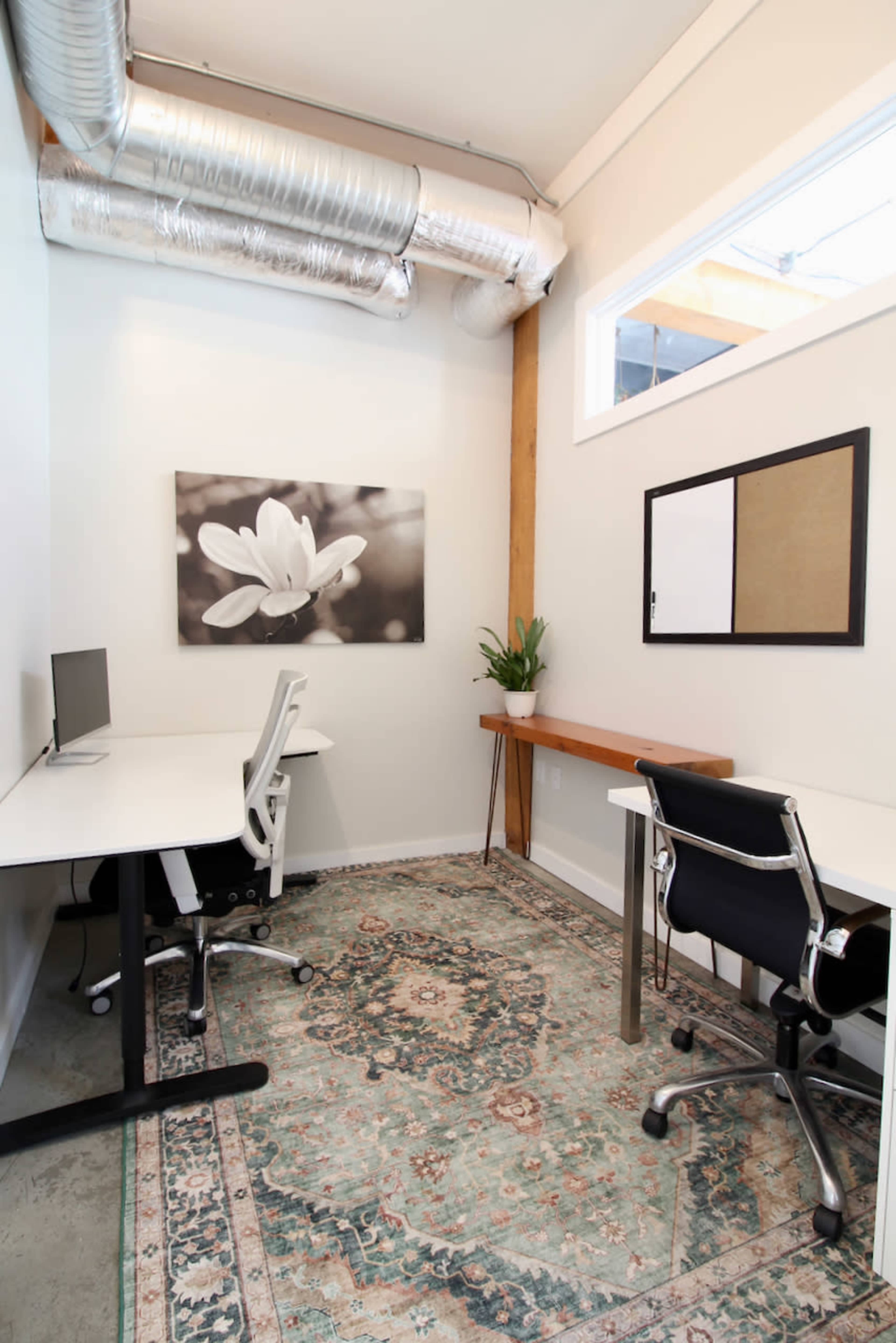 The image shows a small office space with two workstations, a large wall art piece featuring a flower, and a wooden desk along one side against a light-colored wall.