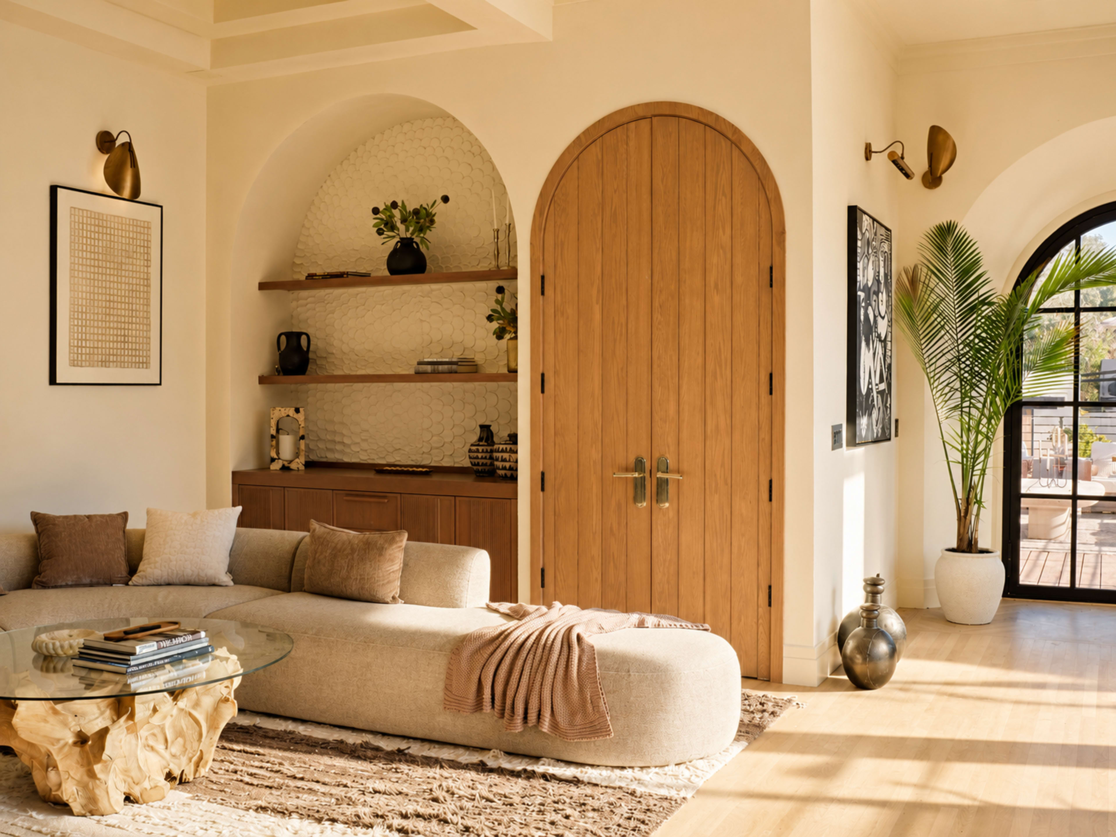 A modern living room features a curved sofa, a round glass coffee table, and a large wooden doorway, complemented by indoor plants and decorative shelves.