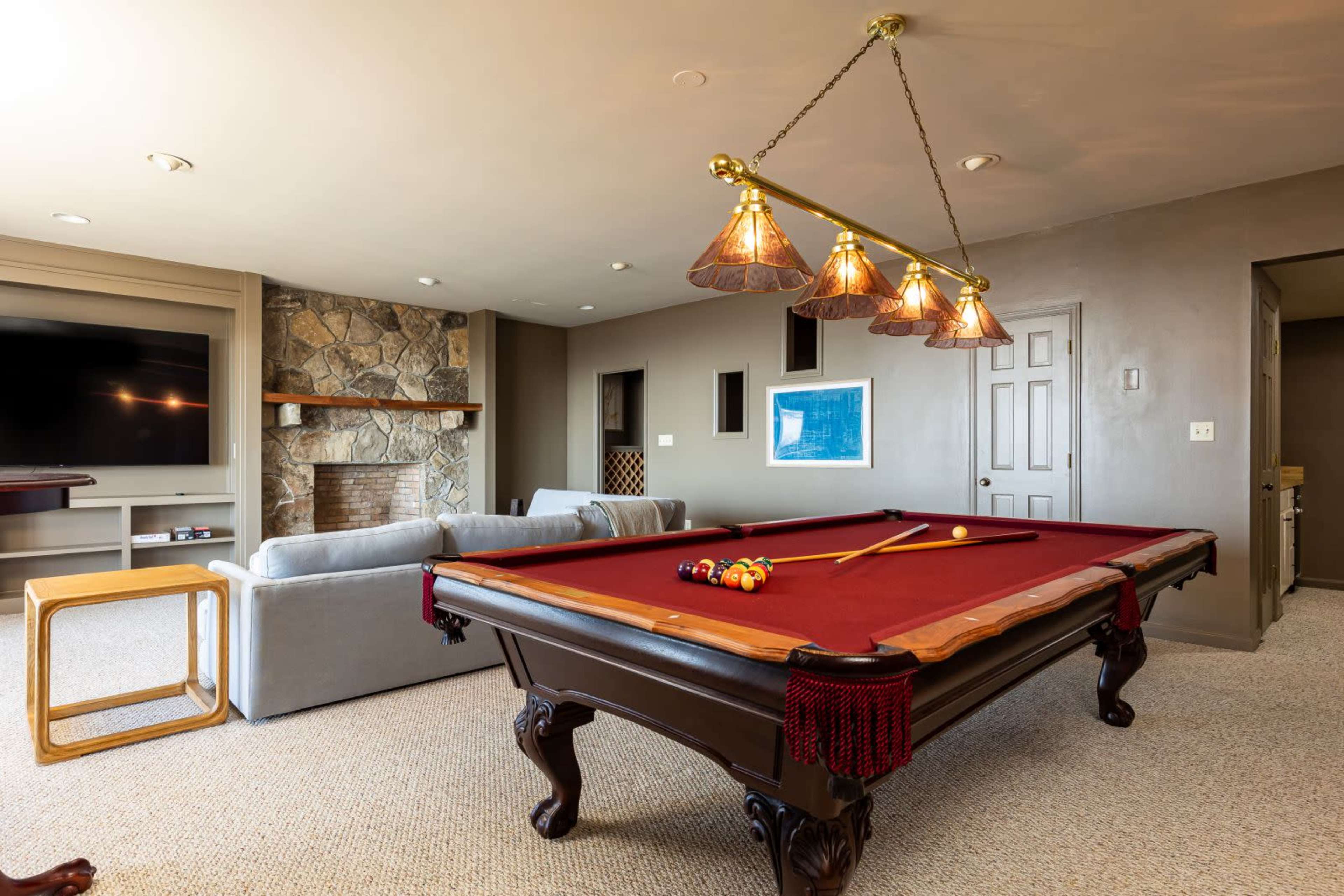 A billiards table with a red felt surface is centrally placed in a room featuring a stone fireplace and a comfortable seating area.