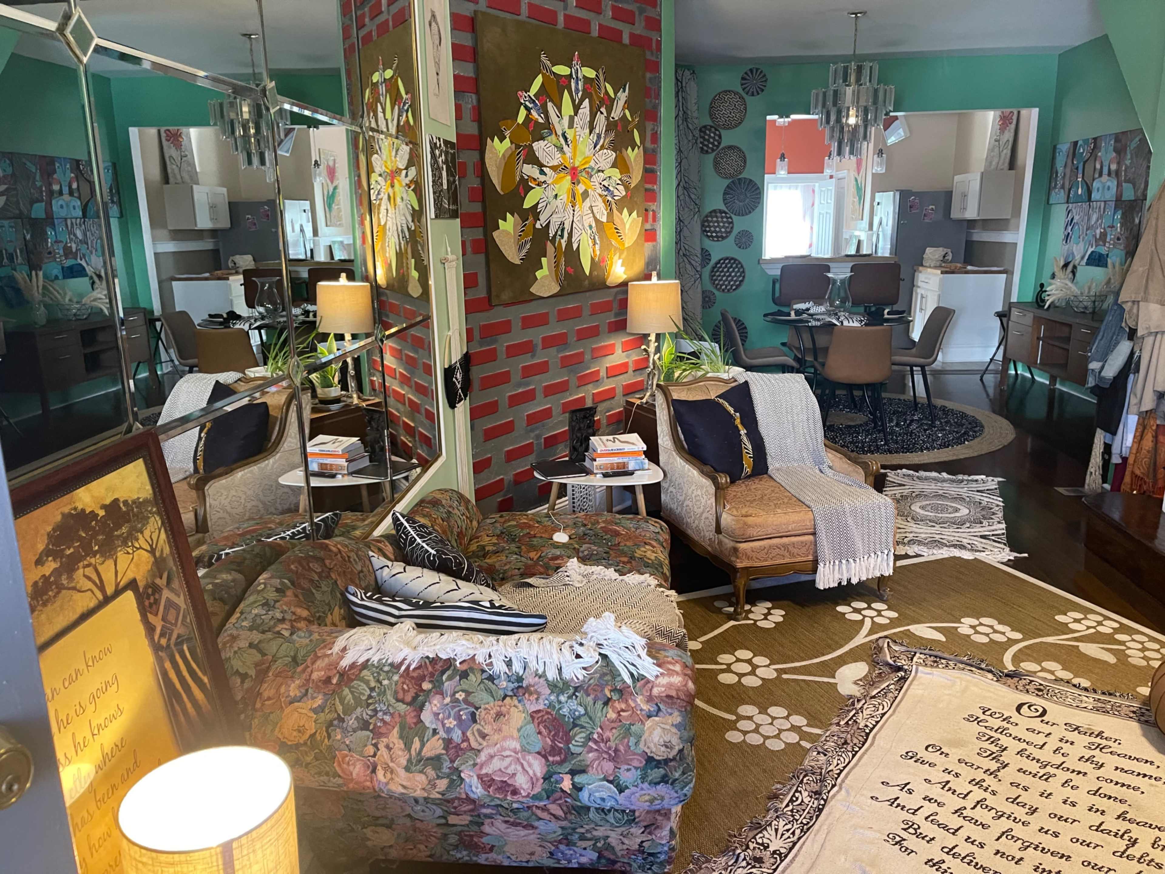 The image depicts a vibrant, eclectic living space featuring a floral-patterned sofa, a coffee table with books, and a mirrored wall reflecting a dining area with modern furniture.