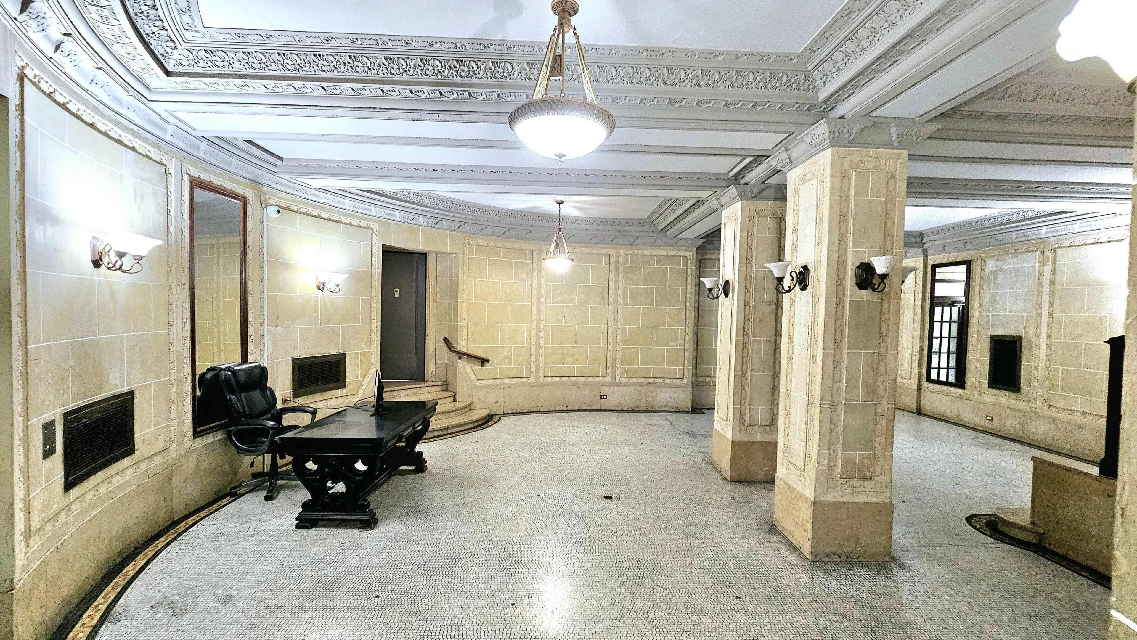 The image shows a spacious, vintage lobby with decorative walls, a dark table, and a pair of chairs, illuminated by a chandelier.
